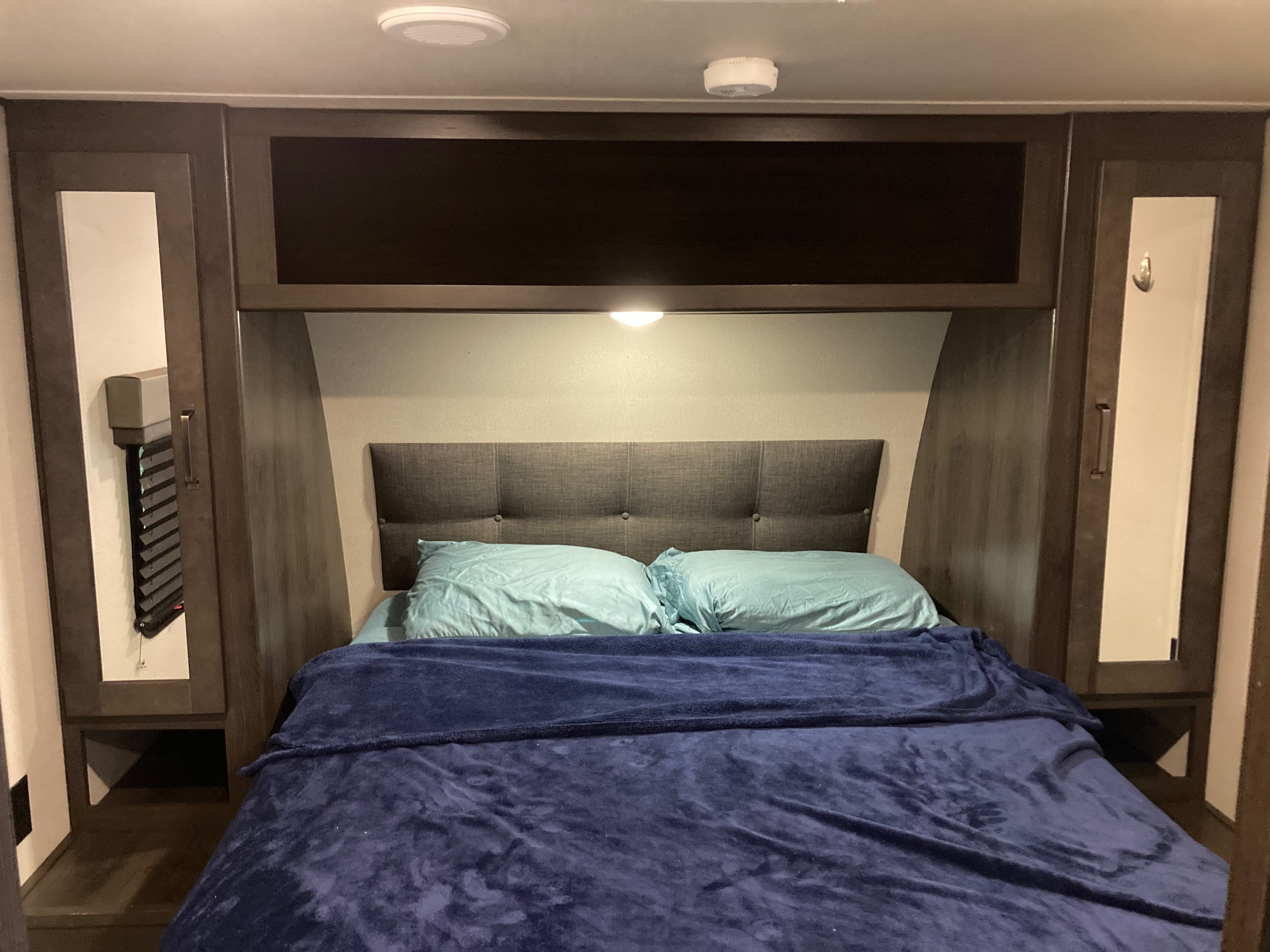 RV rental bedroom with a neat bed, blue bedding, padded headboard, and wooden storage cabinets overhead and beside.. Grand Design Transcend 2021