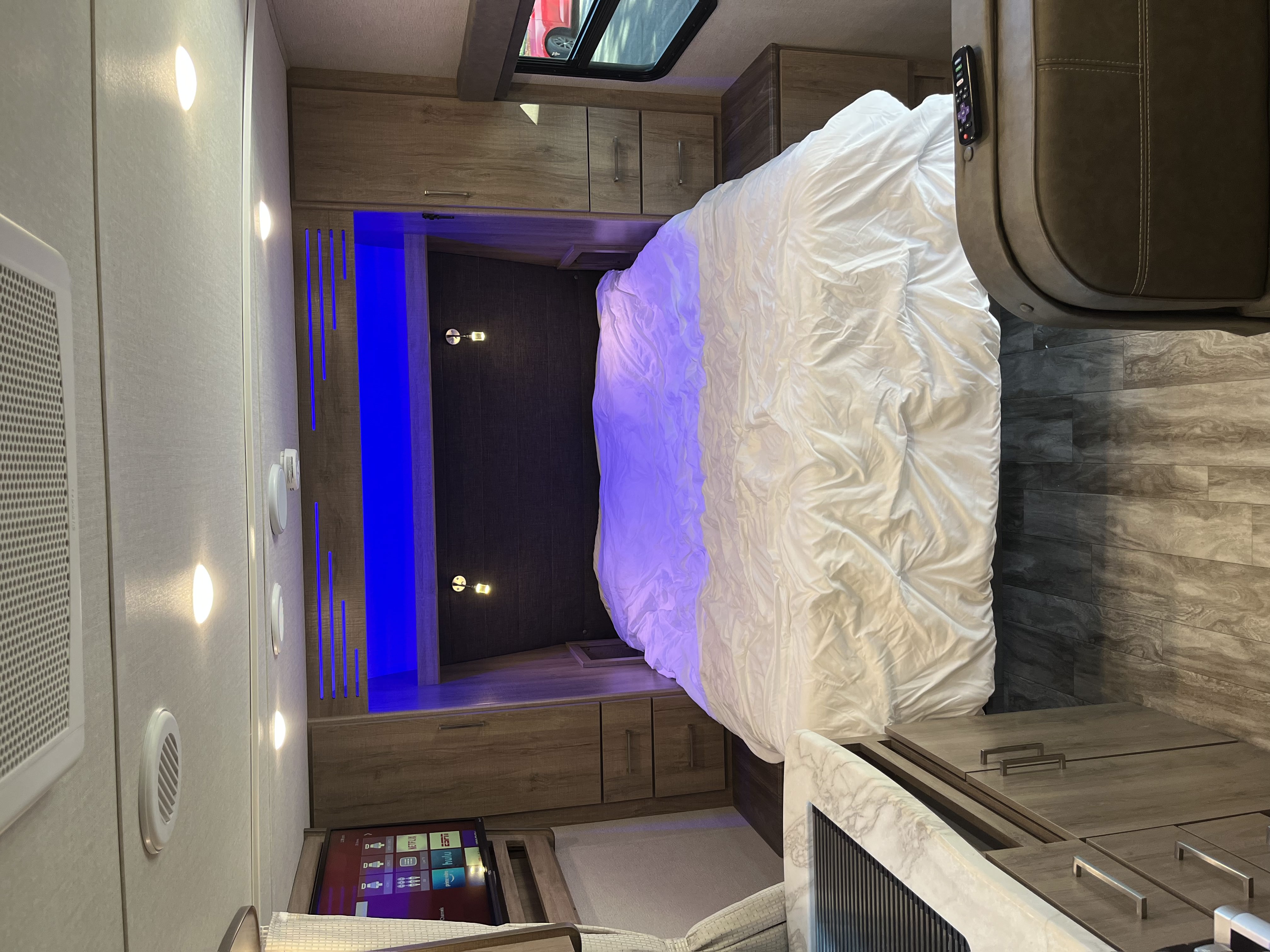 A modern RV rental bedroom offers comfort with a cozy bed, stylish wood accents, and striking blue ambient lighting.. Grand Design Imagine XLS 2022