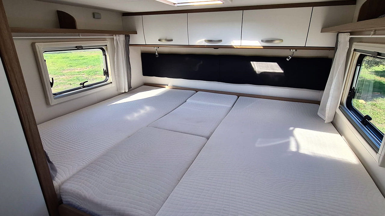 An inviting RV rental bedroom featuring a large bed with segmented white mattresses, overhead cabinets, and bright windows.. Carado T338 2022