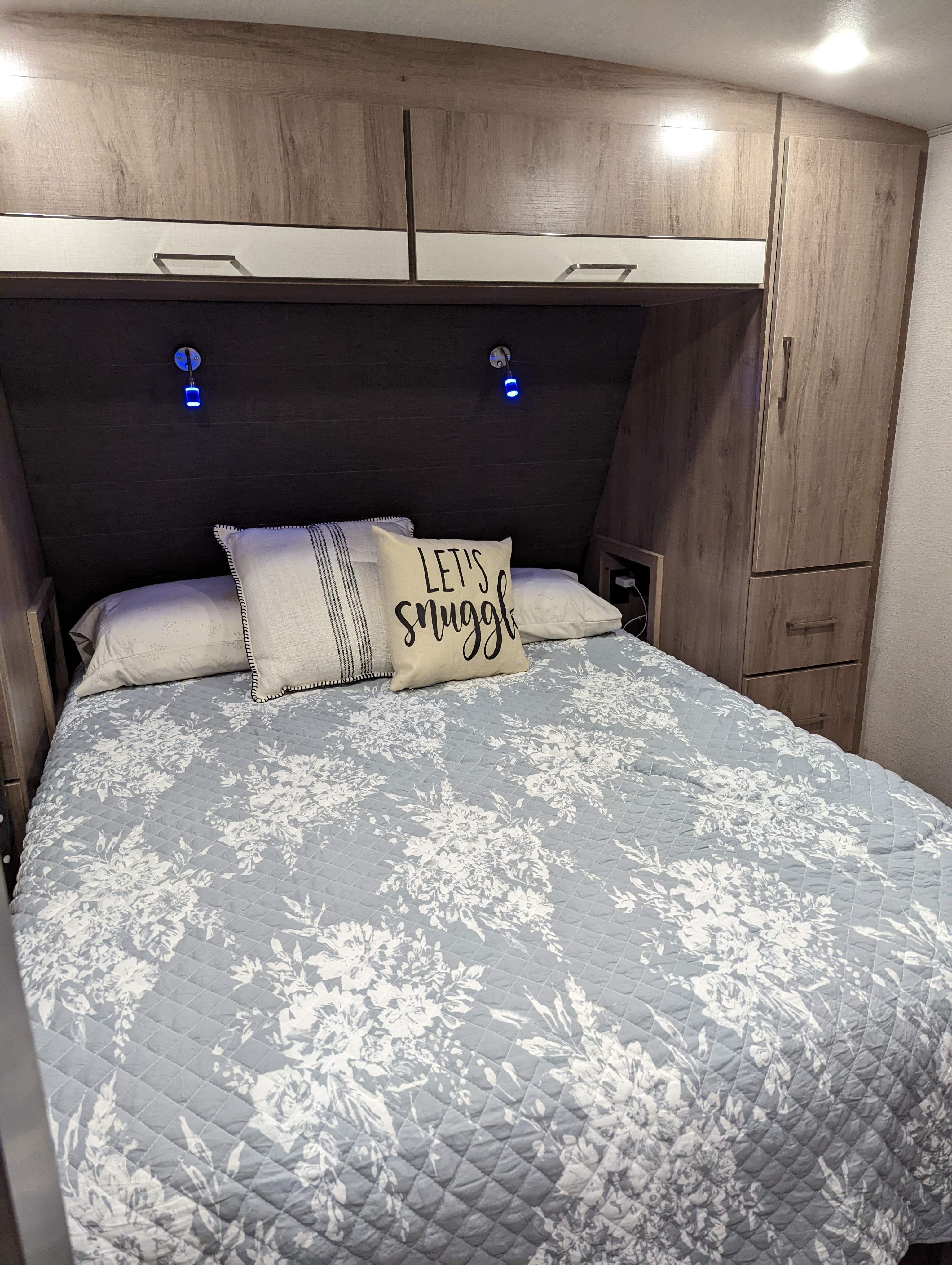 Stay cozy in this RV rental bedroom featuring a blue floral quilt, "snuggle" pillow, built-in cabinets, and reading lights.. Grand Design Imagine 2022