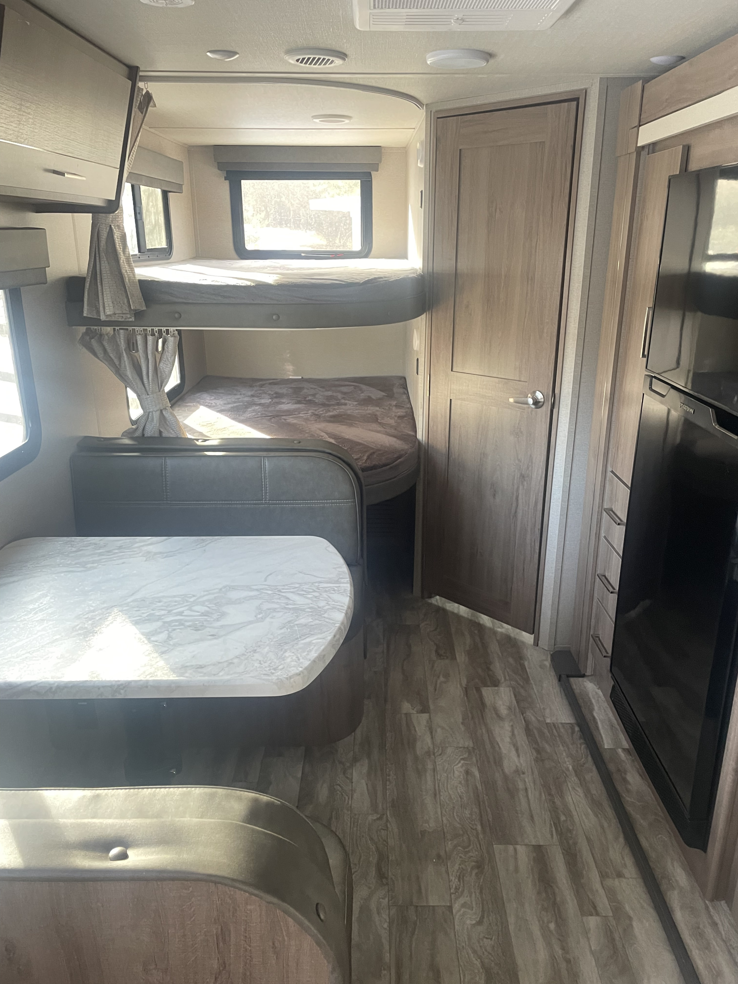 An RV rental's interior shows bunk beds, a dinette with a light marble-look table, refrigerator, and wood-patterned flooring.. Grand design Imagine XLS 2023