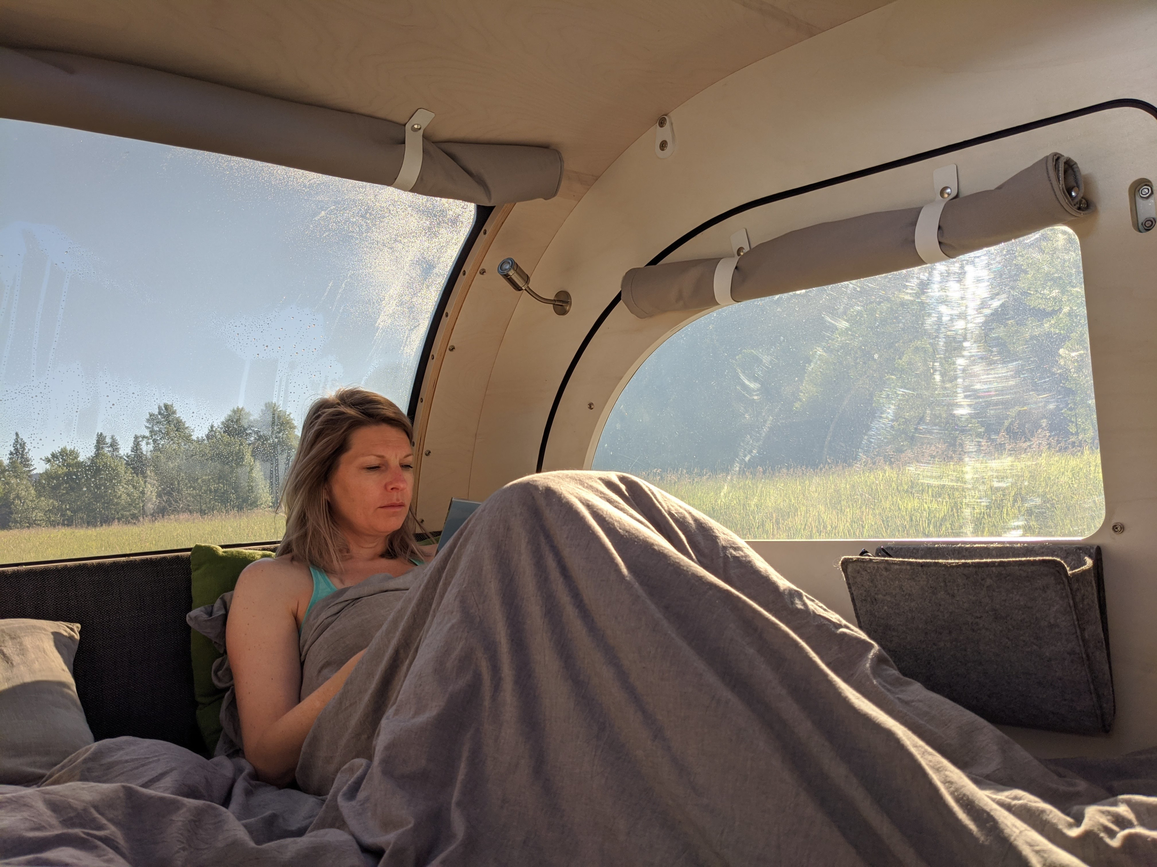 Inside a cozy RV rental: a woman under a blanket, reading with a serene view of trees and fields through the large window.. droplet XL 2023