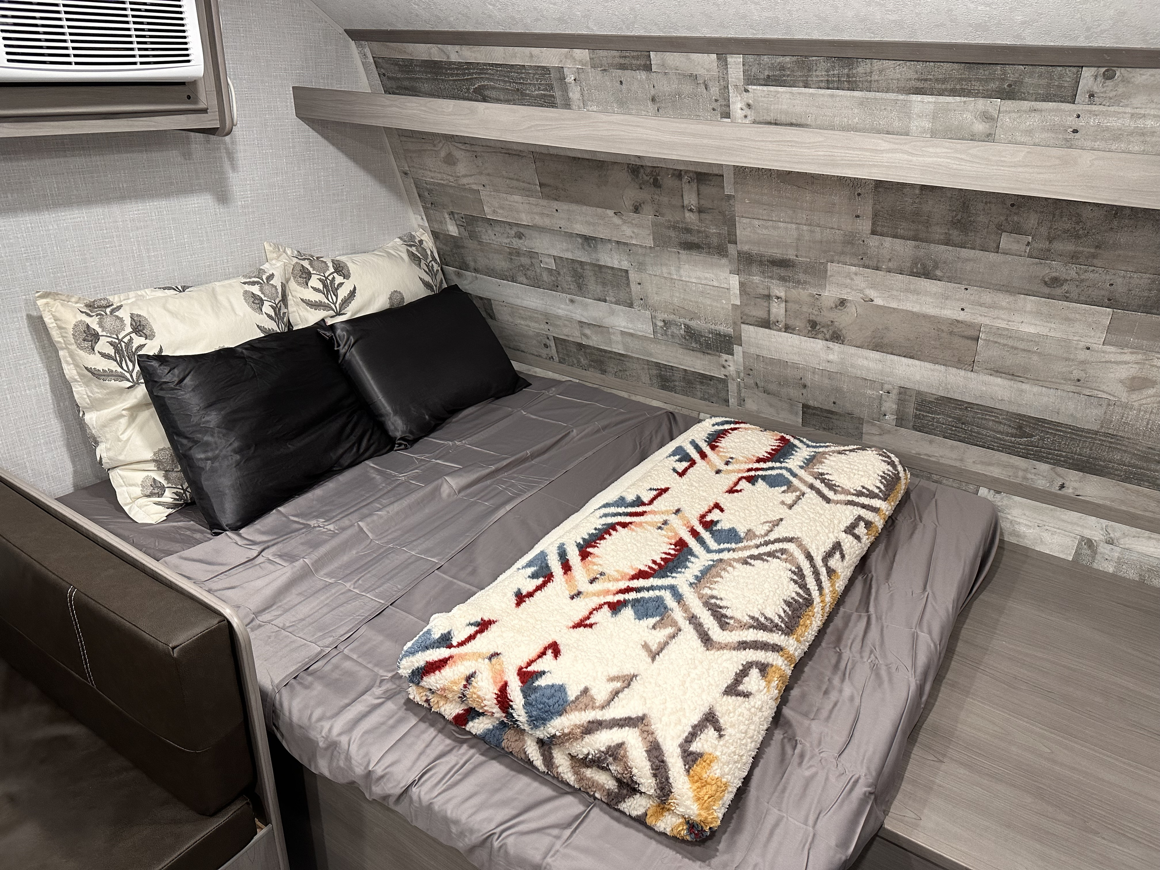 Cozy RV rental bedroom features a bed with patterned and black pillows, a soft blanket, and rustic wood-paneled walls.. Coleman LANTERN LT 17B 2023