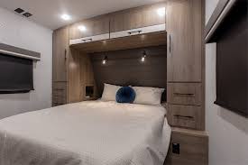 An inviting RV rental bedroom. Features a queen bed with white bedding, wooden built-ins, and reading lights, perfect for travel.. Grand Design Imagine 2021