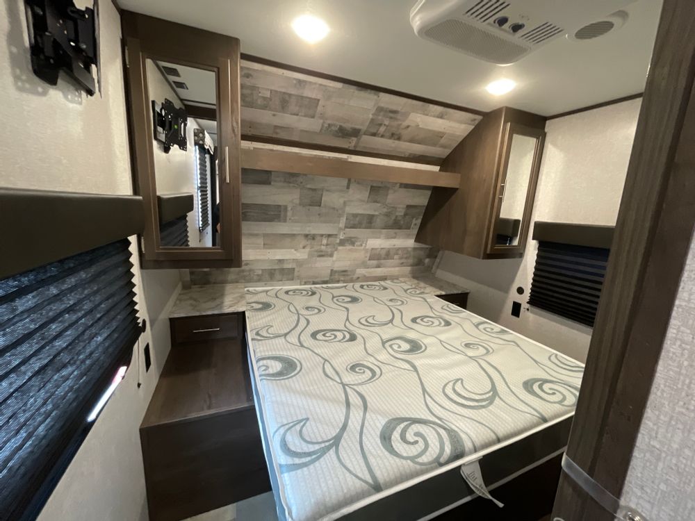 Step into this RV rental bedroom with a patterned mattress, overhead cabinets, wall-mounted TV bracket, and accent wall.. Dutchmen Coleman Lantern 2020