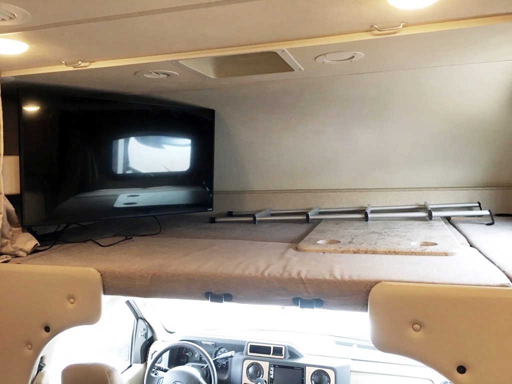 Inside an RV rental: the over-cab bunk bed area with a mounted TV, a ladder, and a portable table with cup holders.. Thor Chateau 2018
