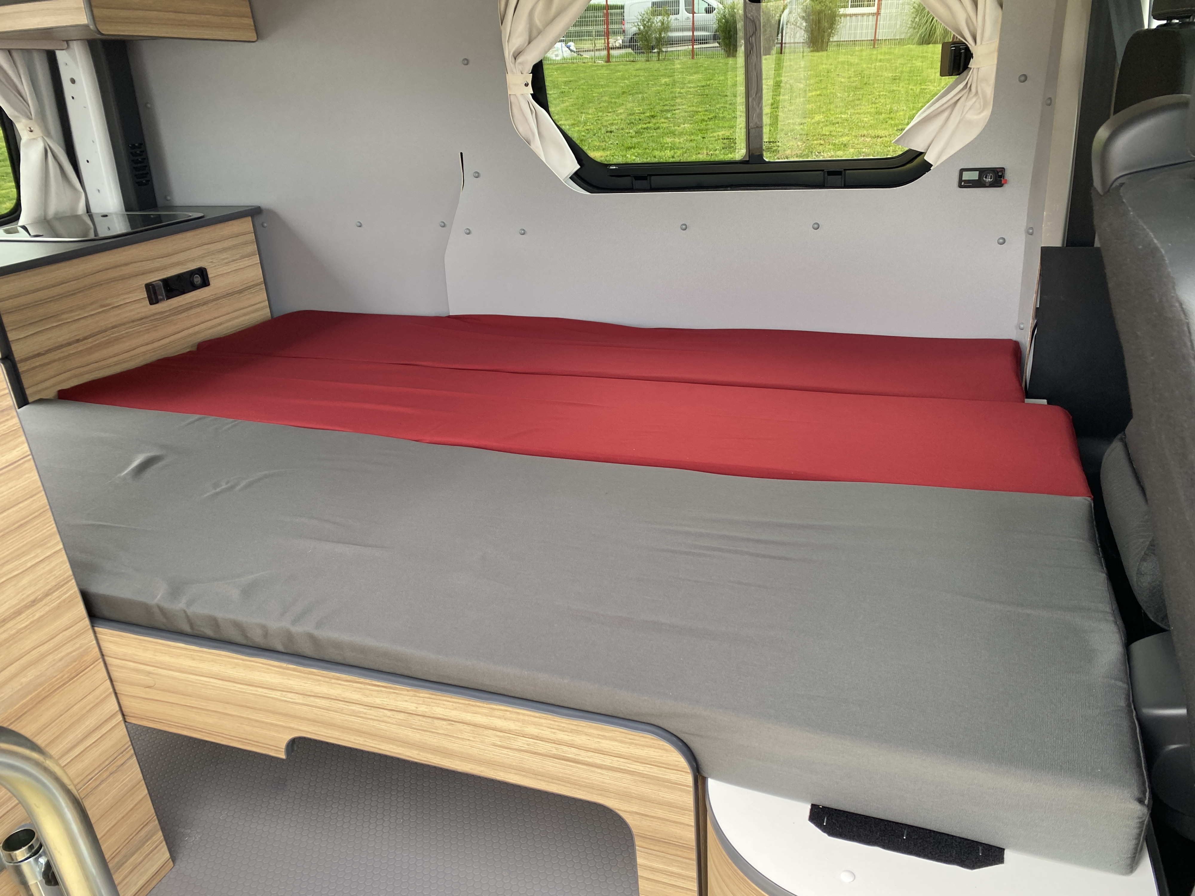 RV rental interior showcasing a cozy, convertible bed with grey and red cushions, wooden cabinetry, and windows, ideal for travel.. Renault TRAFIC 2023