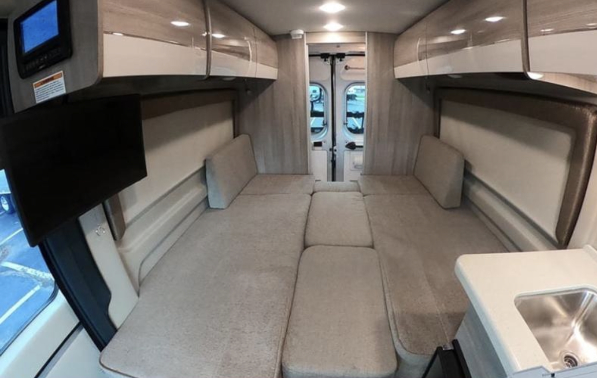 RV rental interior view: A beige cushioned bed dominates the space, with a TV, sink, and overhead cabinets.. Thor Sequence 2021