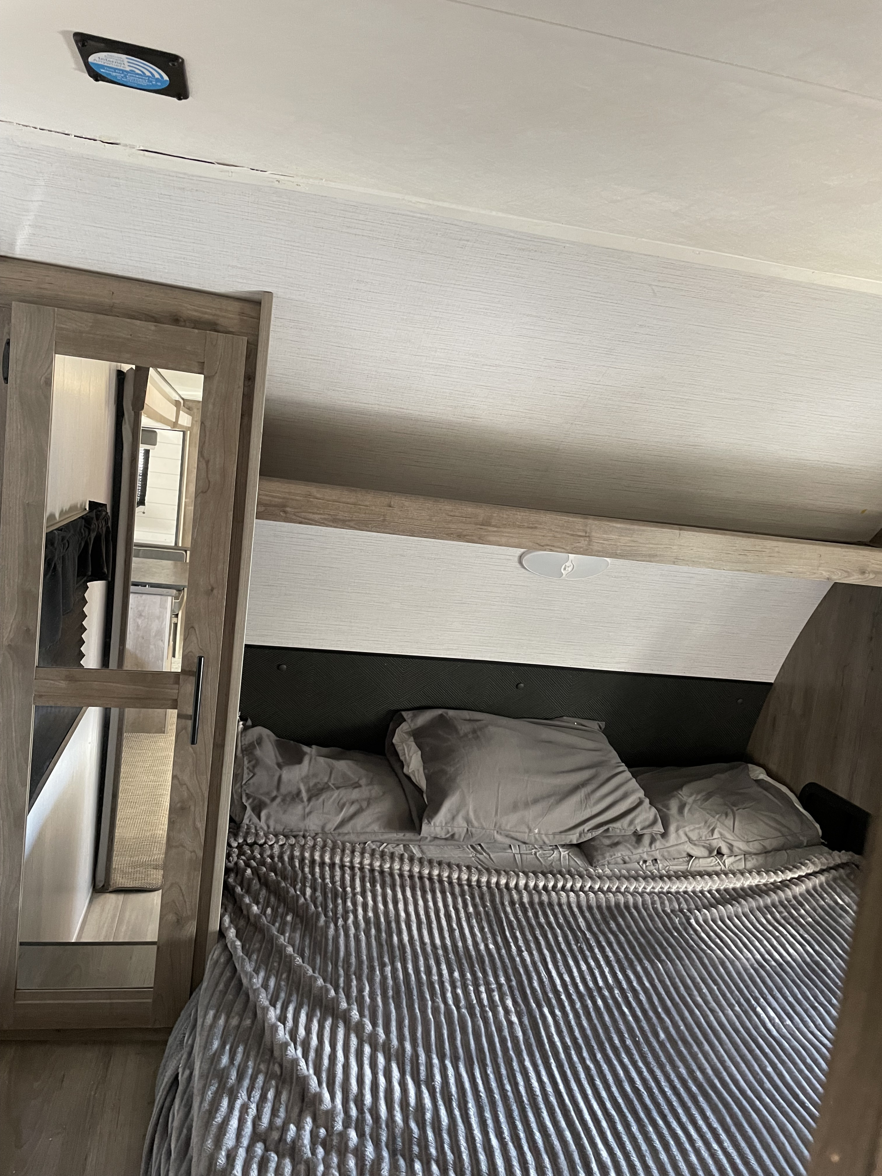 Inside an RV rental bedroom, showcasing a comfortable bed with grey pillows and a textured blanket, wood-look walls, and ceiling fixtures.. Heartland RVs Pioneer 2021