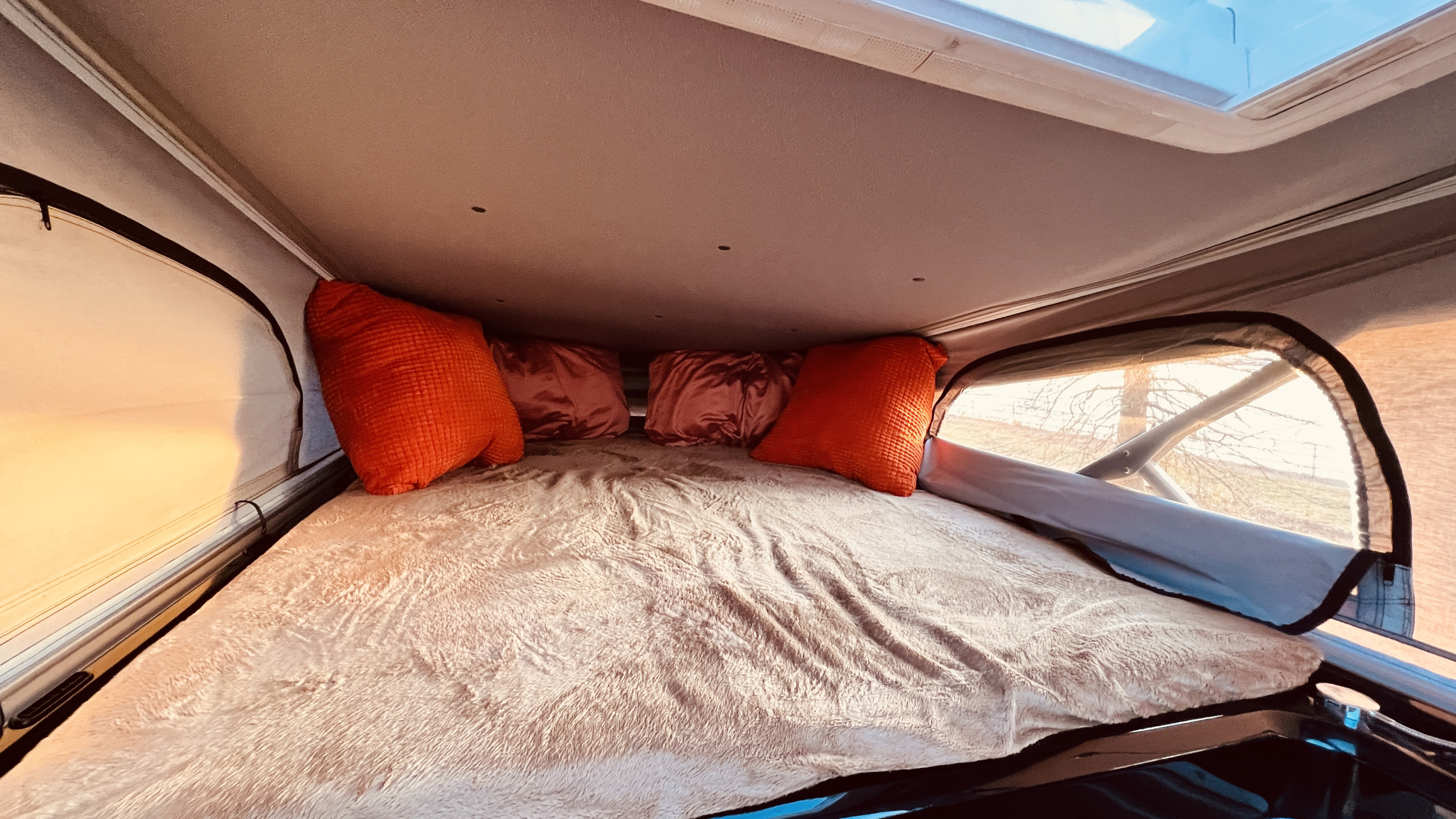 Cozy pop-top sleeping area in an RV rental, featuring a plush bed, colorful pillows, and natural light from overhead and side windows.. Thor Sequence 2022