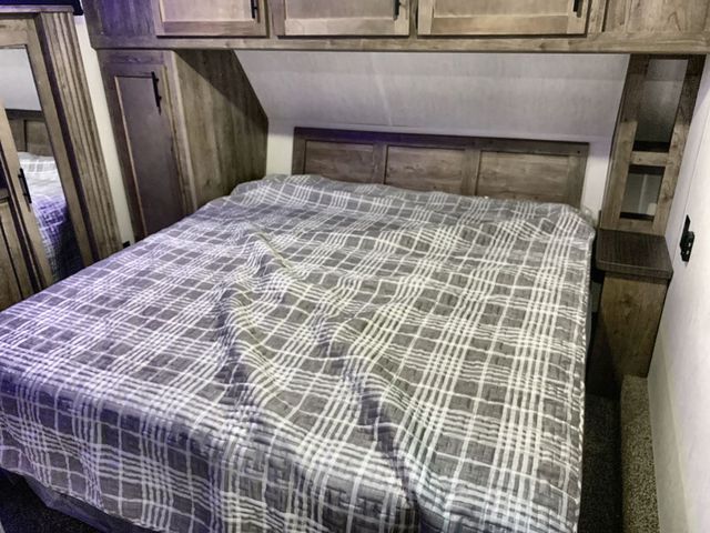 Cozy RV rental bedroom features a comfy plaid bed, wooden headboard, and overhead storage cabinets.. Forest River Wolf Pack 2022