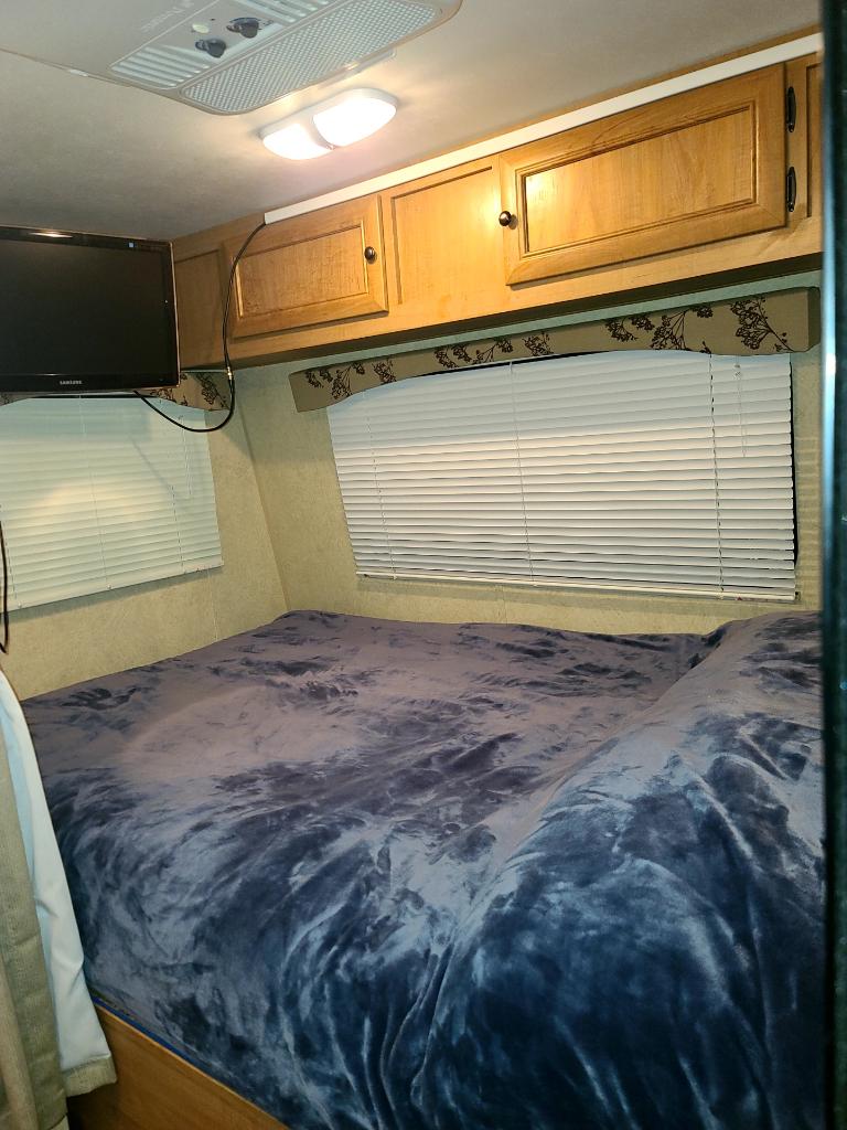 Inside this RV rental, find a comfortable bedroom featuring a plush blue blanket, overhead wooden cabinets, TV, and window blinds.. Nomad 173 2013