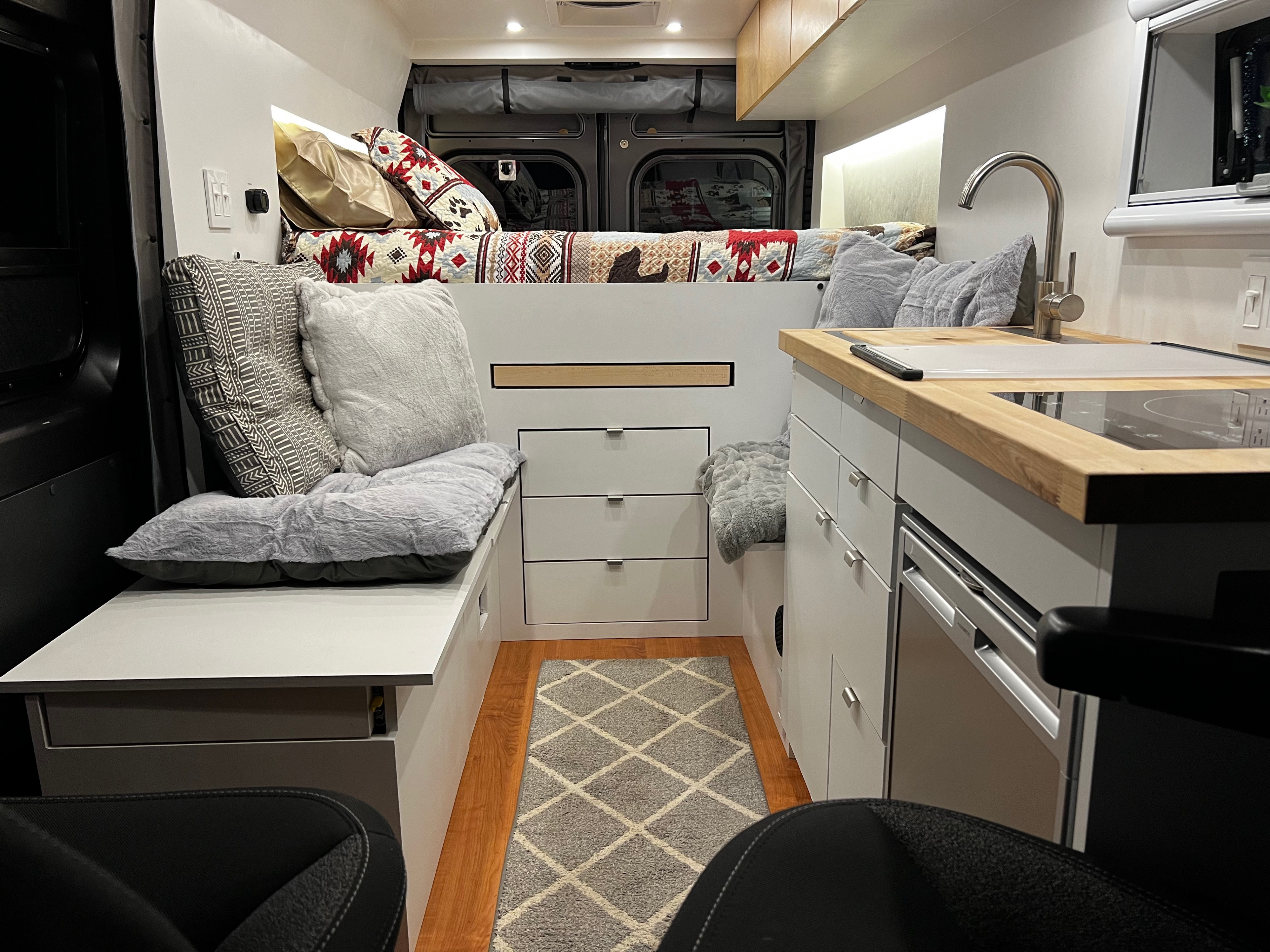 Inside a comfy rv rental, featuring a bed with patterned quilt, compact kitchen, and a plush seating area with storage.. Dodge Ram Promaster 3500 2021