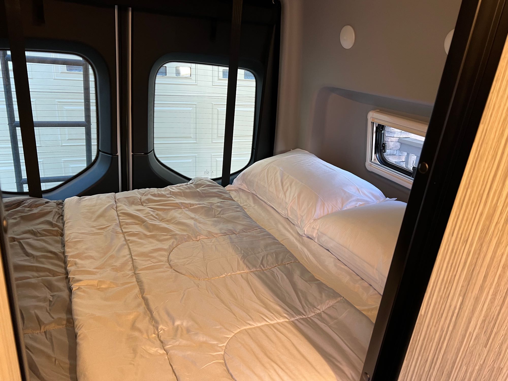 A neatly made bed with light-colored bedding fills the back of an RV rental, featuring windows on the side and rear.. Winnebago Revel 2020