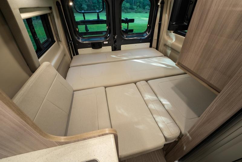 RV rental's versatile interior: a light-colored bed/seating area, with rear windows overlooking a green field.. Thor Motor Coach Sequence 20J 2023