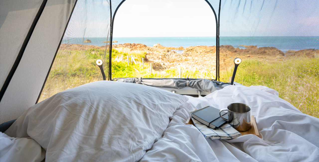 RV rental: Cozy bed with white linens, mug, and notebook, facing a stunning rocky coastline and ocean view through a mesh tent.. Bürstner T600 2024