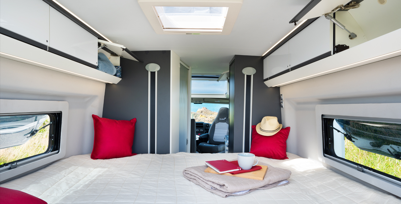Inside an RV rental: a bright, cozy bedroom with a large bed, overhead storage, and scenic window views for comfortable travel.. Bürstner T600 2024