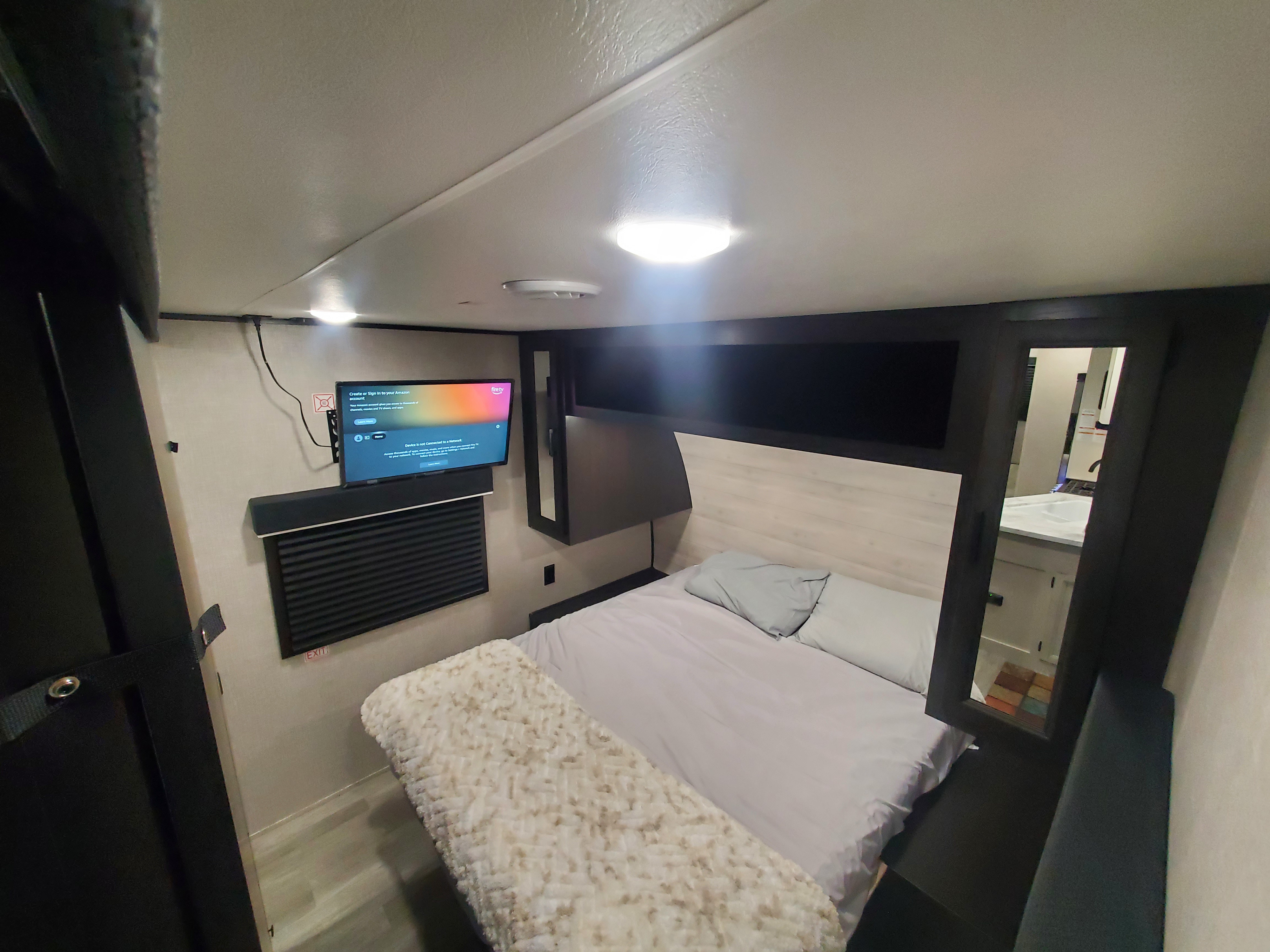 Cozy RV rental bedroom with wall-mounted TV & soundbar. Features a comfortable bed, overhead lighting, and an "EXIT" sign visible.. Jayco Jay Flight 2023