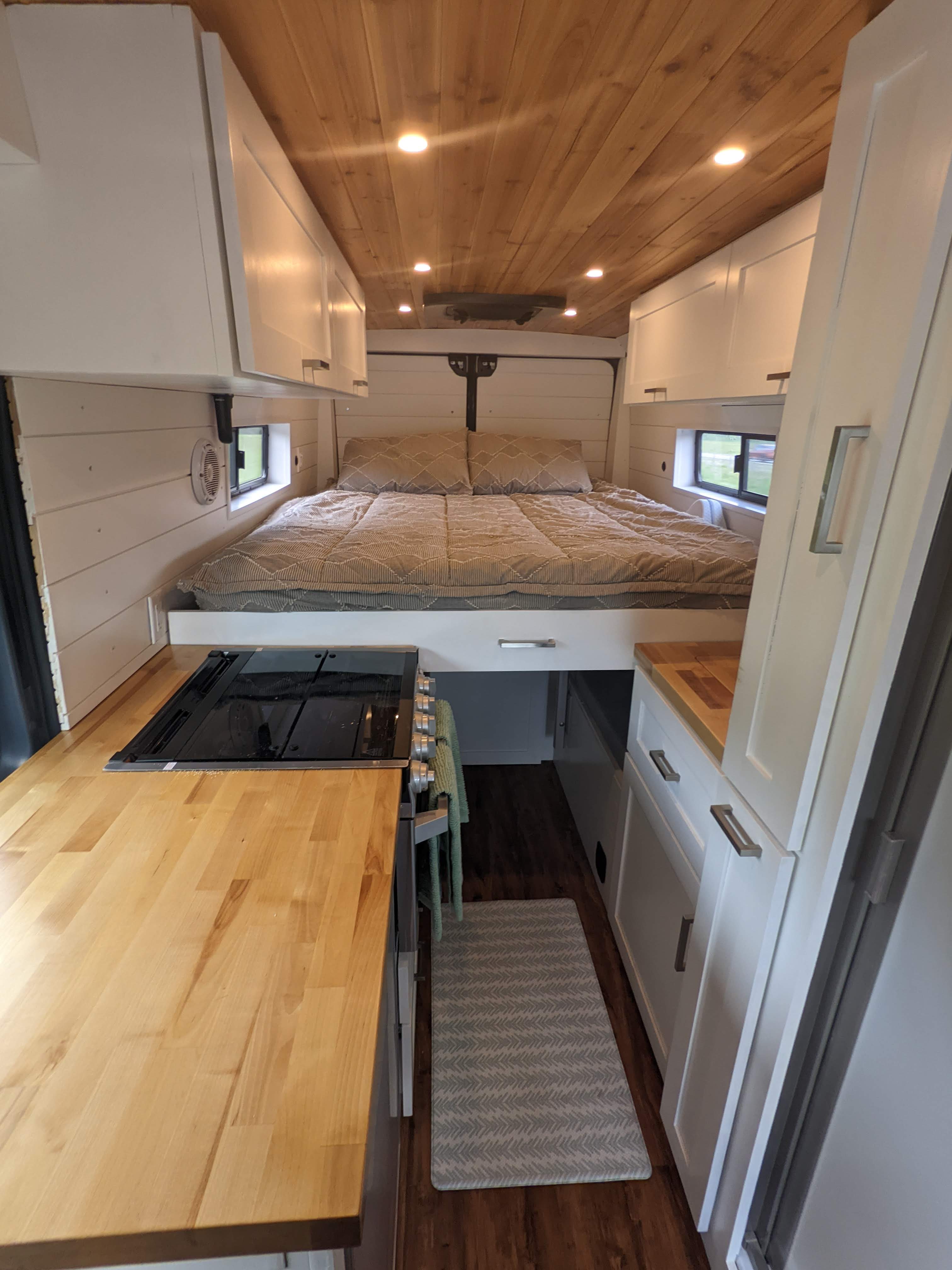 Cozy RV rental interior shows a compact bed, integrated kitchen, warm wood ceiling, and white storage cabinets.. RAM Promaster 2019