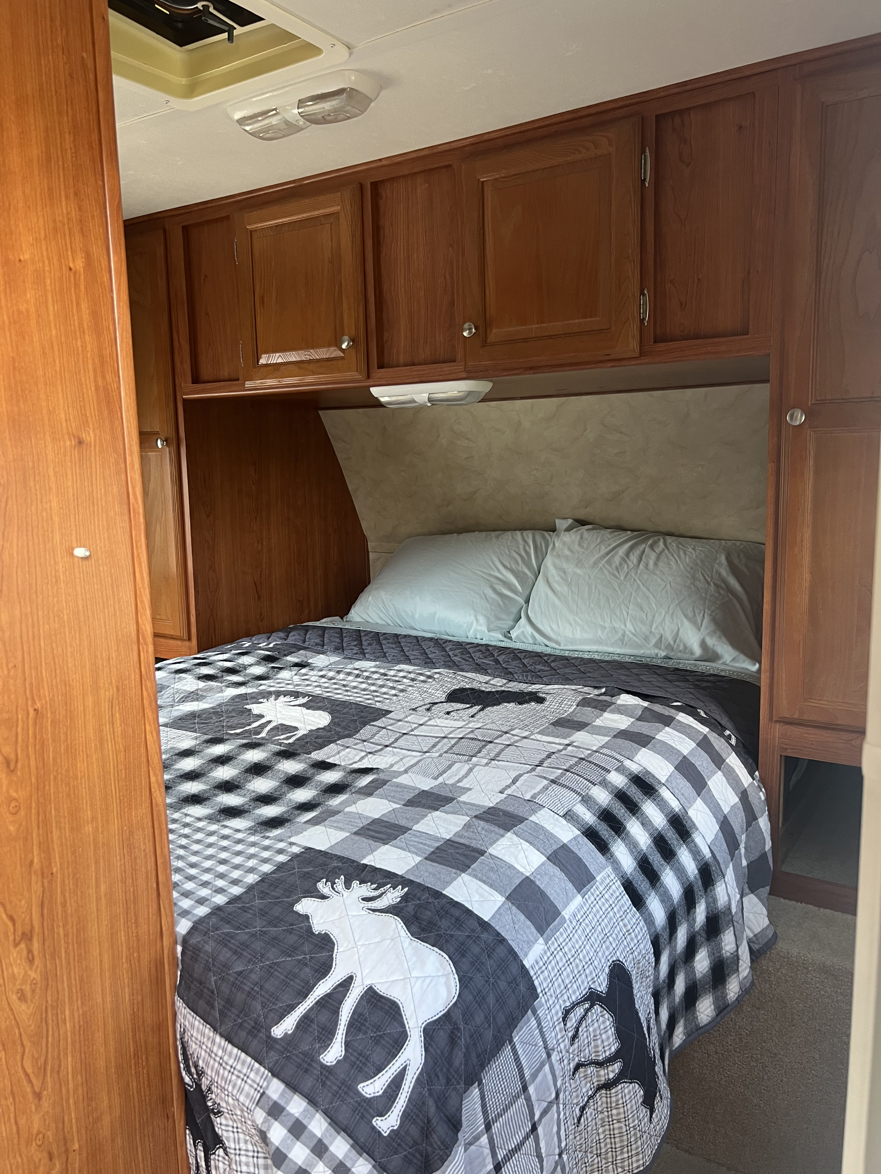 Enjoy comfort in this RV rental bedroom: bed with moose quilt, wooden cabinets, and bright overhead light.. Holiday Rambler Campmaster 2011