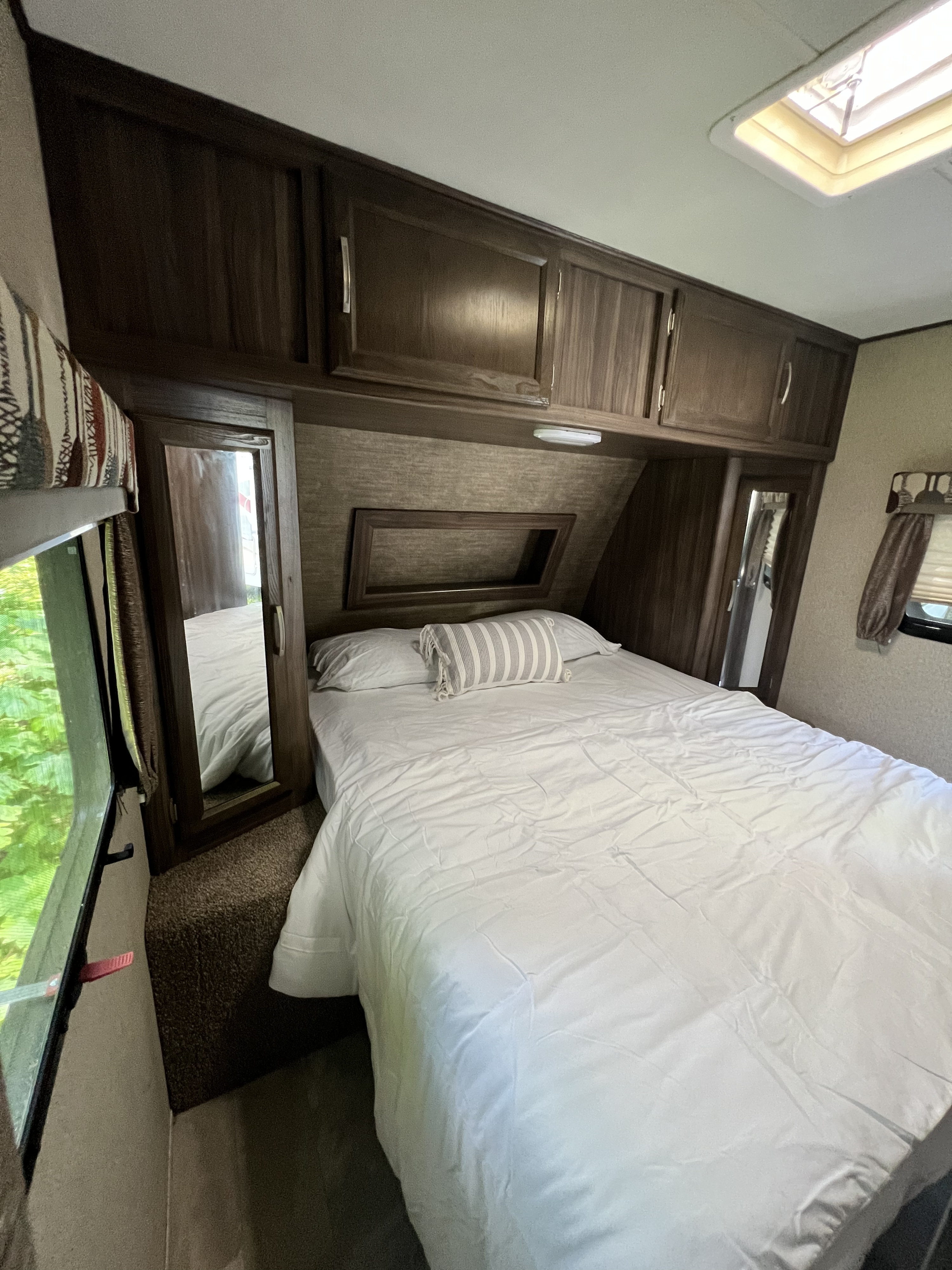 Spacious RV rental bedroom. Features a cozy bed with white linens, wood cabinets, and natural light from a window and skylight.. Coachmen Apex 2018
