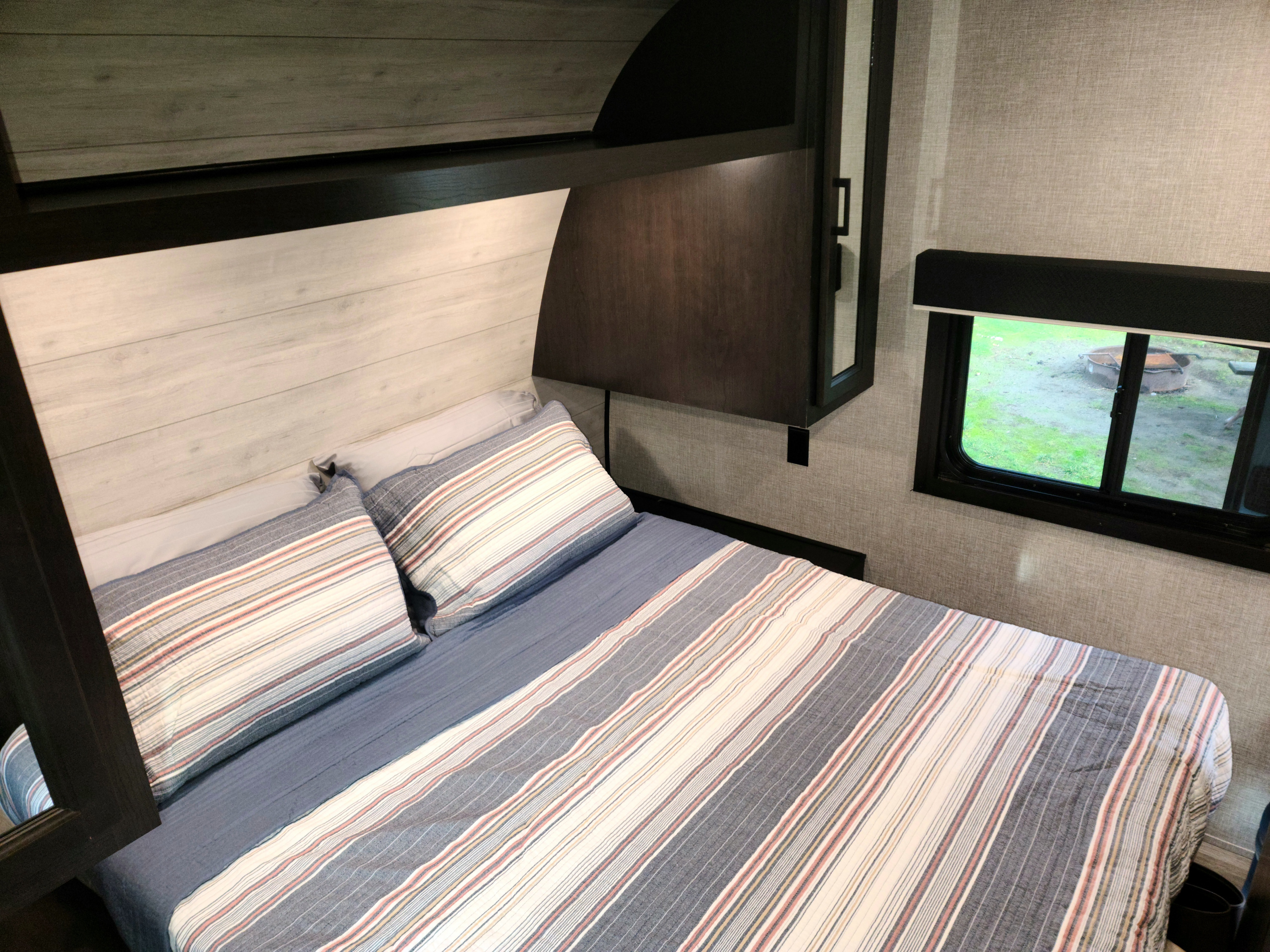 Cozy RV rental bedroom with striped bedding, a window overlooking a yard with a fire pit, and overhead cabinets.. Jayco Jay flight 242BHSW 2023