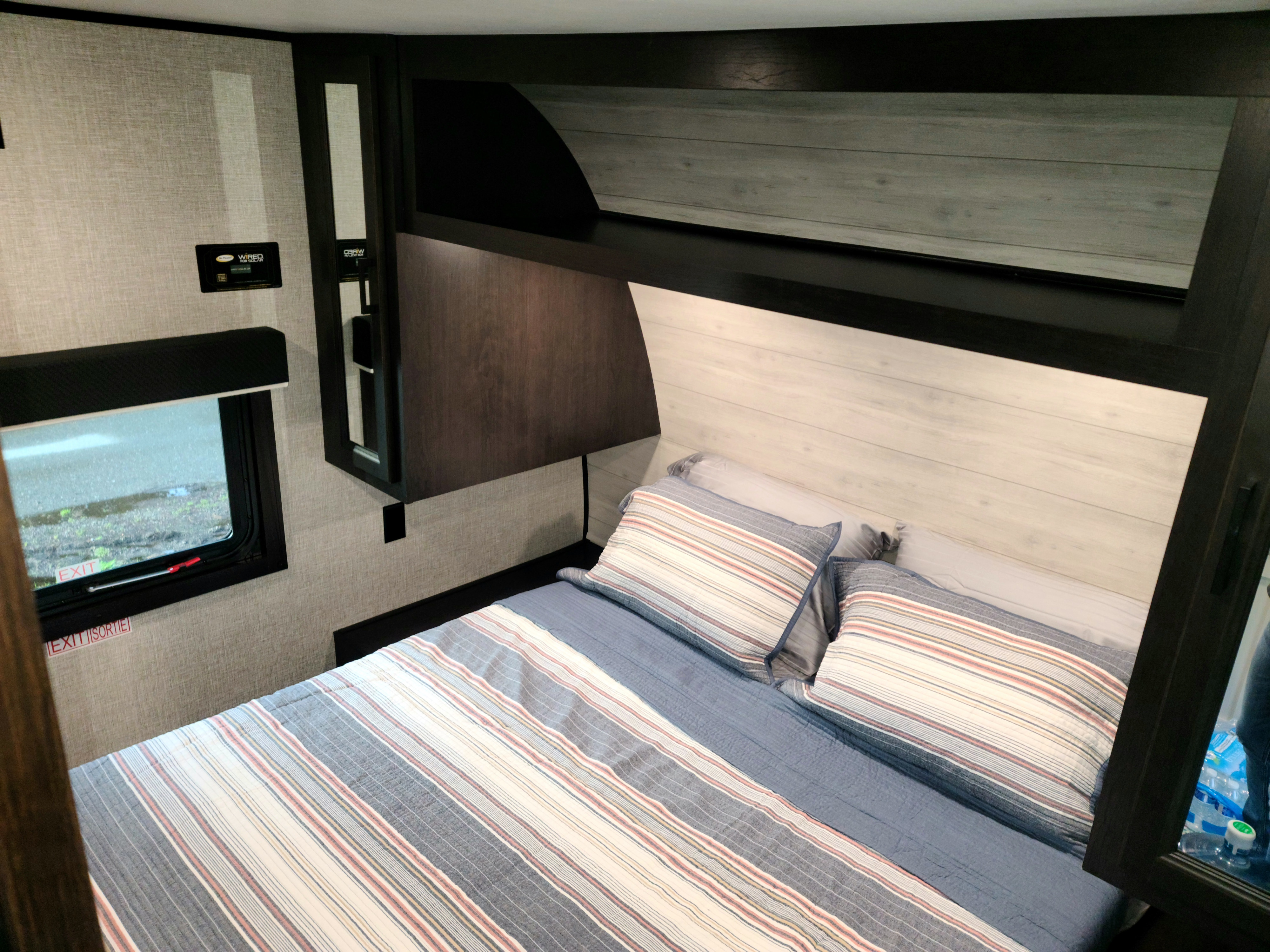 Inside an RV rental: a modern bedroom with striped bedding, overhead storage, and a solar-ready system control panel.. Jayco Jay flight 242BHSW 2023
