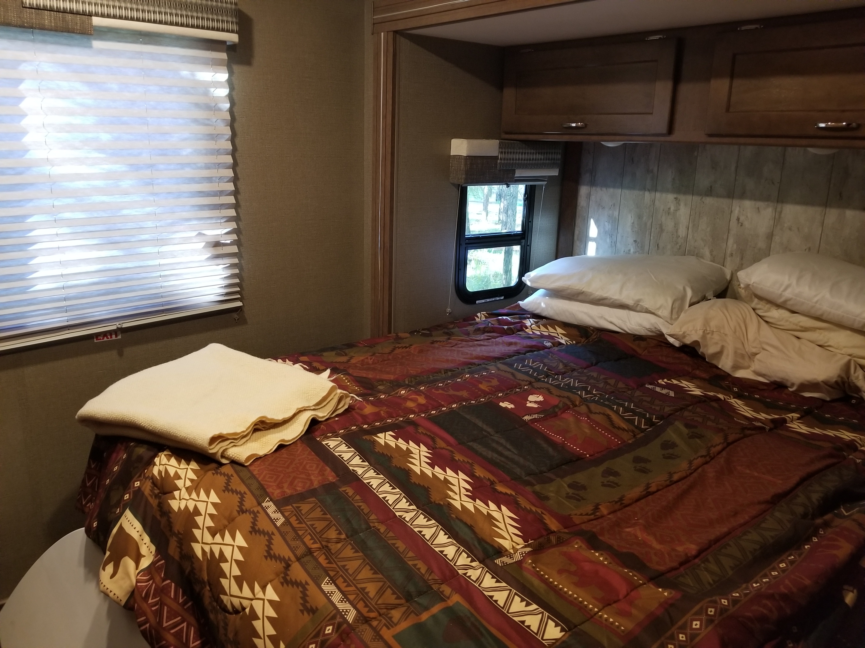 Comfortable RV rental bedroom with patterned comforter, white pillows, and a window with blinds, ready for your next trip.. Winnebago Outlook 2019