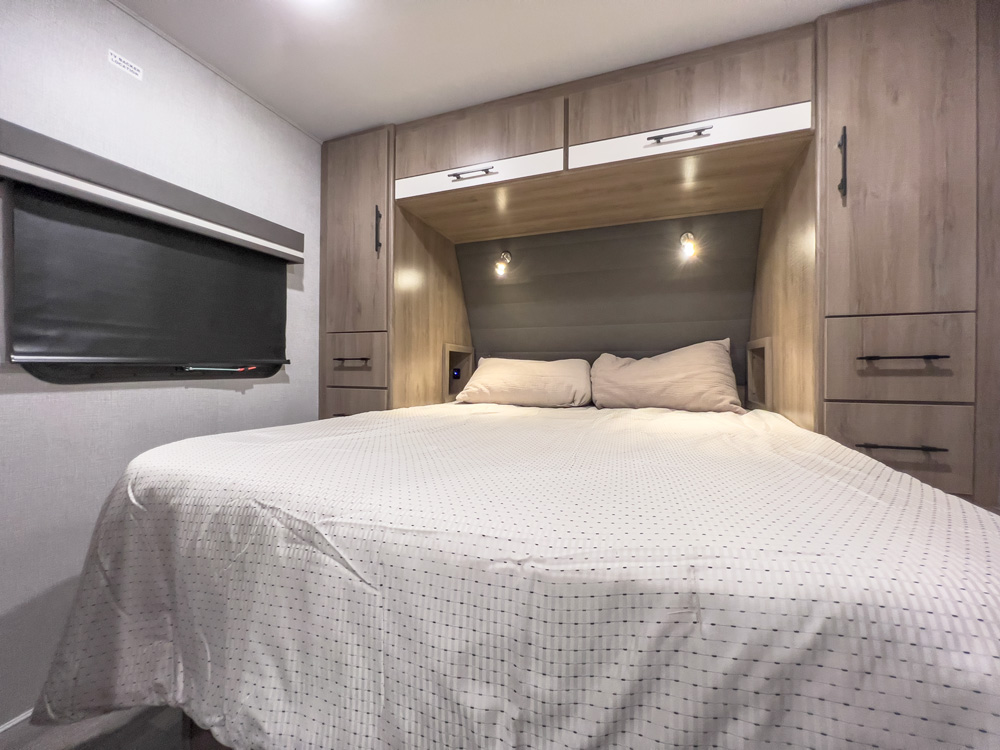 RV rental bedroom with a bed centered between built-in light wood cabinets, a wall-mounted TV left, and reading lights above.. Grand Design Imagine XLS 2022