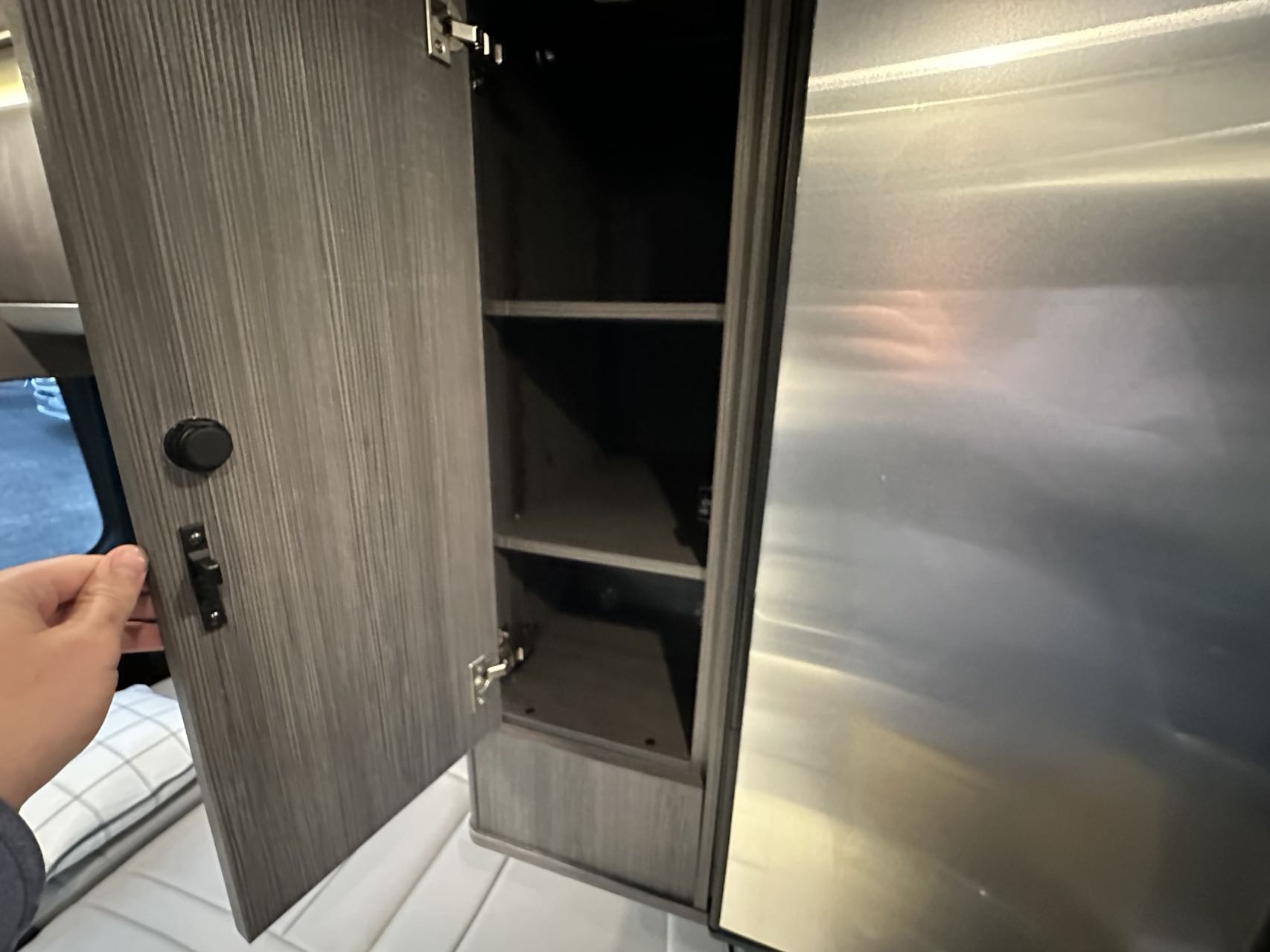 RV rental interior: Hand opens a compact wood-grain cabinet with shelves beside a metal appliance, showcasing smart storage.. Airstream Interstate 2021