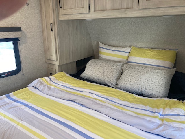 Comfortable RV rental bedroom shows a bed with striped bedding, patterned pillows & wall cabinets.. East to West Della Terra 2022
