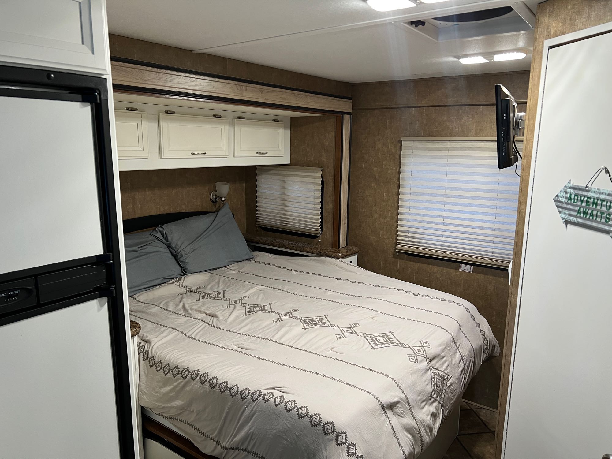 Cozy RV rental bedroom featuring a patterned bed, overhead cabinets, wall-mounted TV, and blinds.. Thor Four Winds 2014