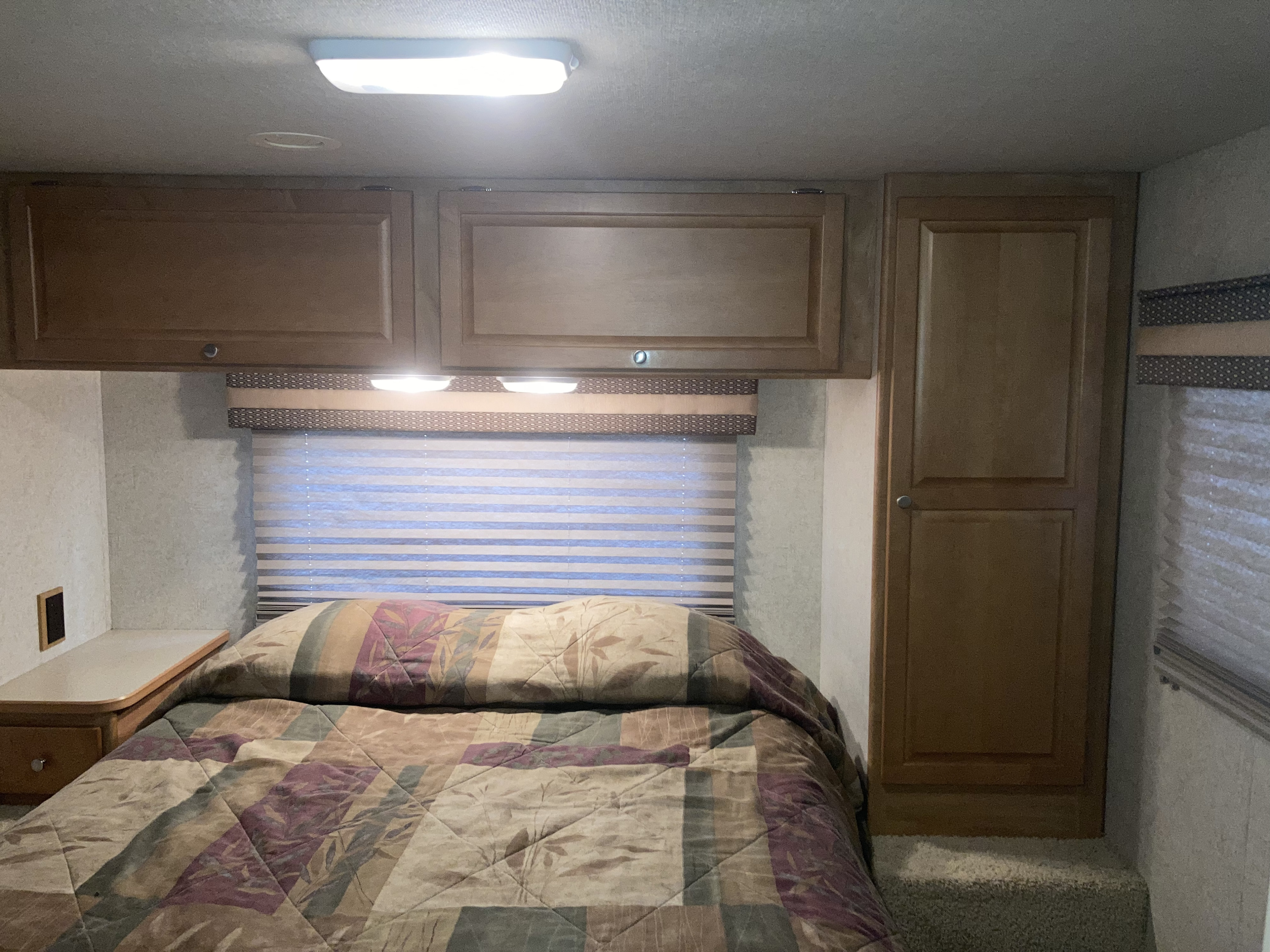 Cozy RV rental bedroom interior with a patterned bed, overhead cabinets, and window blinds, illuminated by bright lights.. Winnebago Chalet 2009