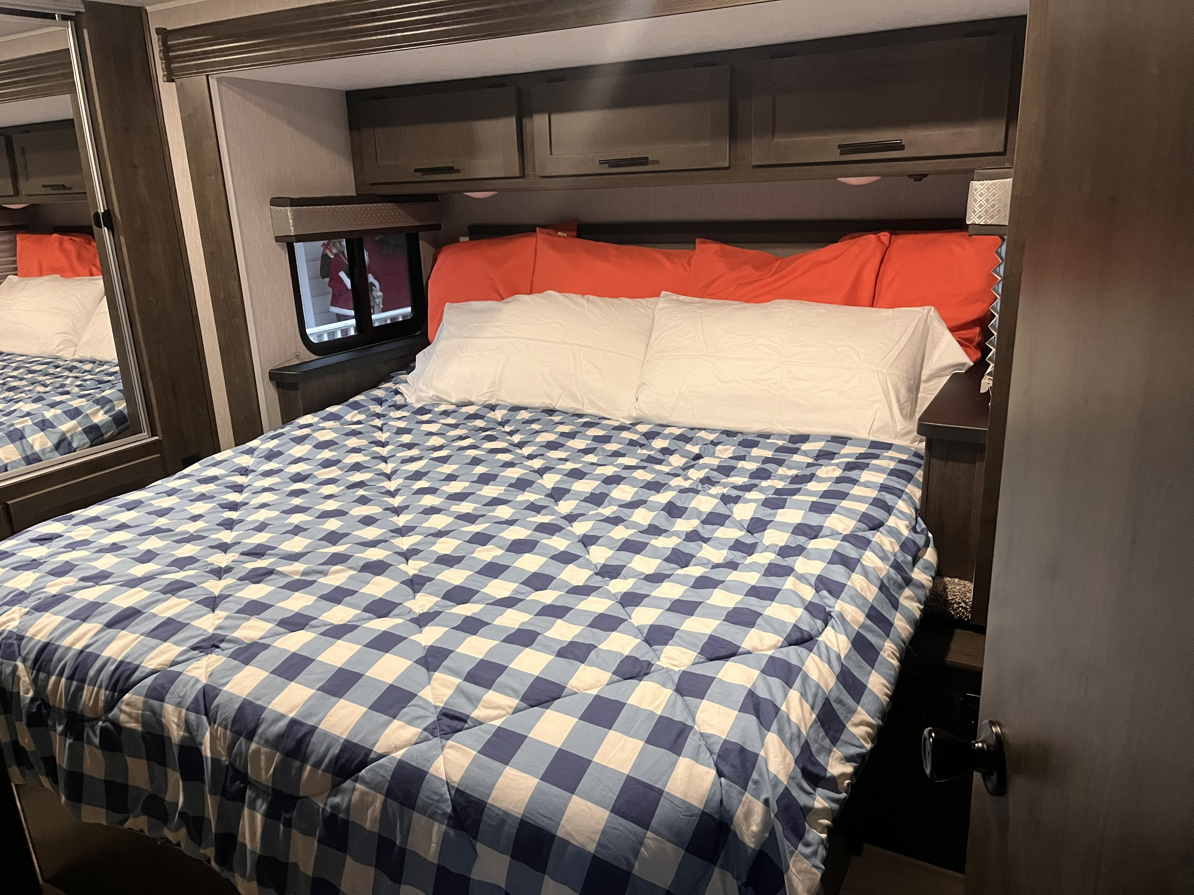 A cozy RV rental bedroom featuring a bed with a blue and white checkered comforter, orange and white pillows, and overhead cabinets.. Coachmen Leprechaun 2023