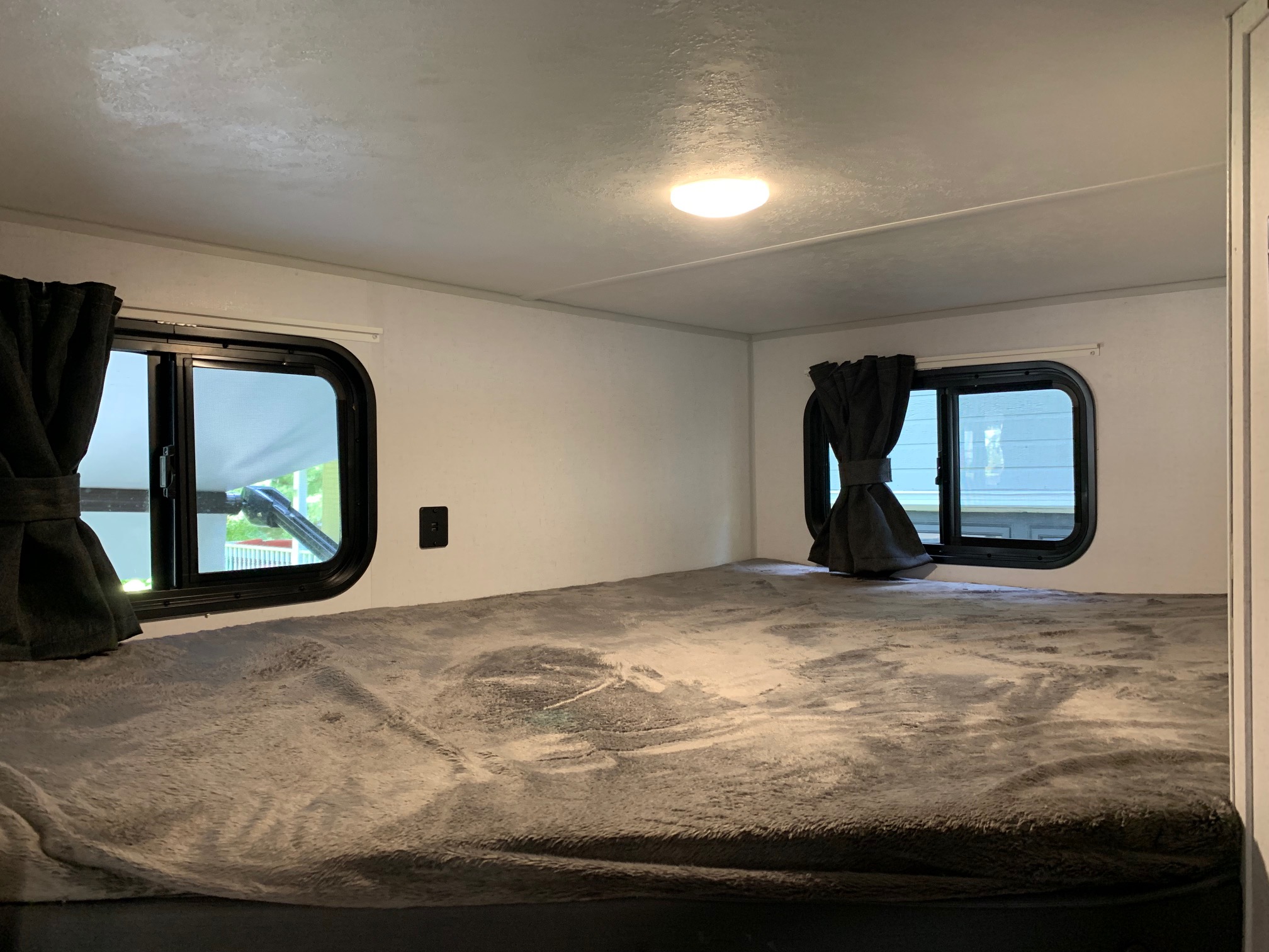Cozy RV rental sleeping area featuring a wide bed with a soft grey blanket, two windows with dark curtains, and an overhead light.. Forest River Salem FSX 2021