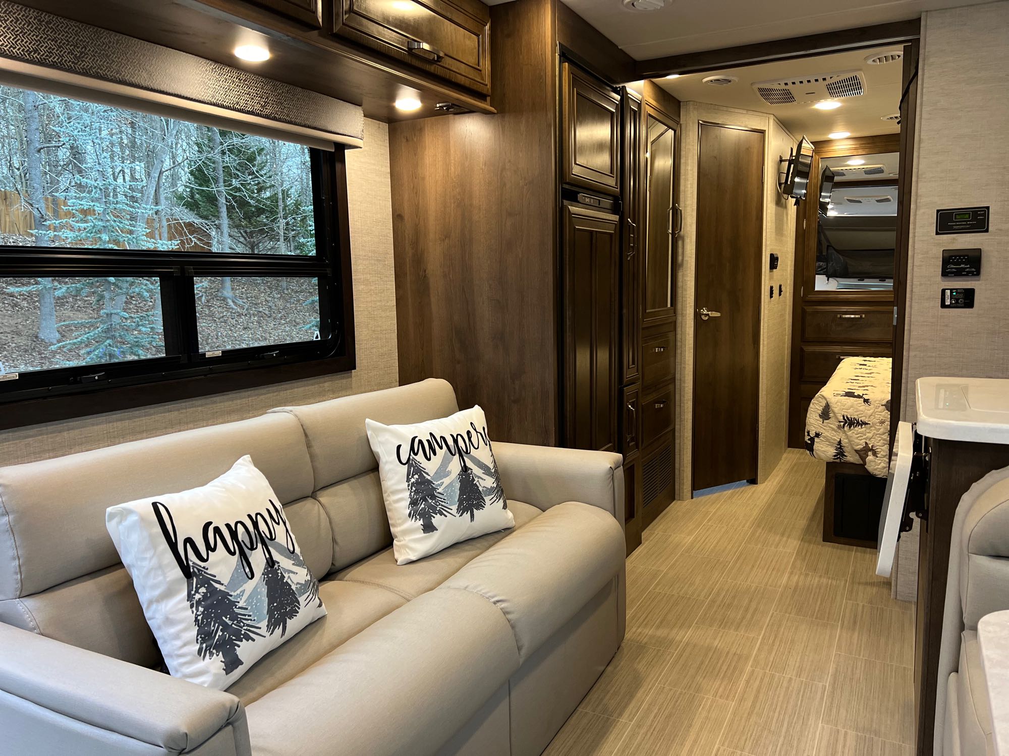 Bright RV rental interior showing a beige sofa, window with tree view, and dark wood cabinetry.. Entegra Coach Esteem 27U 2022