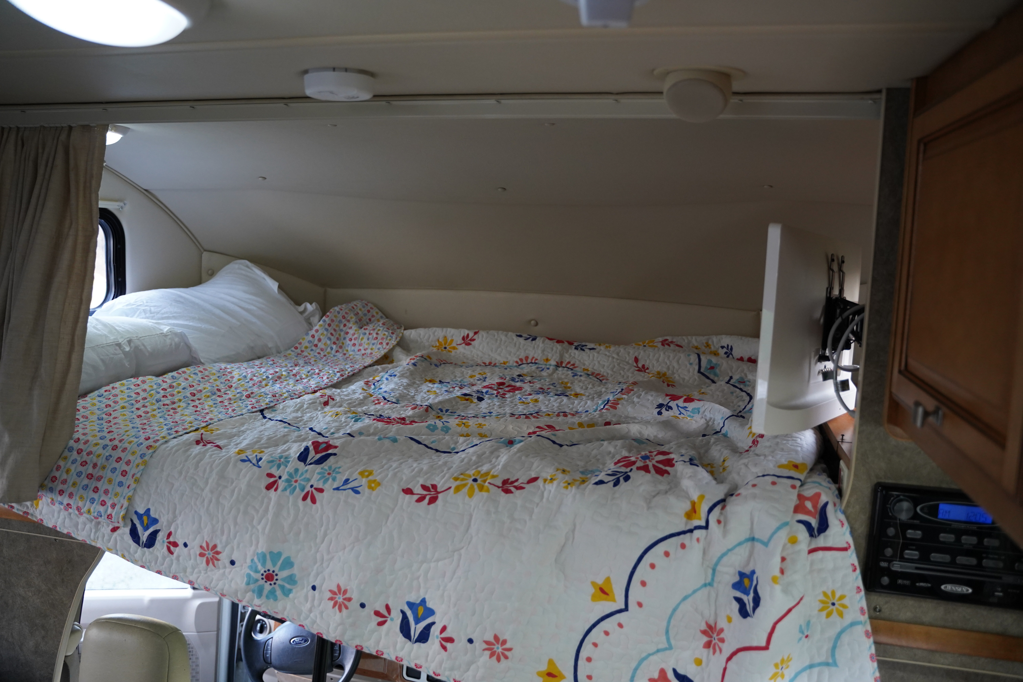 Cozy RV rental cab-over bed with floral quilt and curtained window, ready for travel.. Itasca Impulse 2013