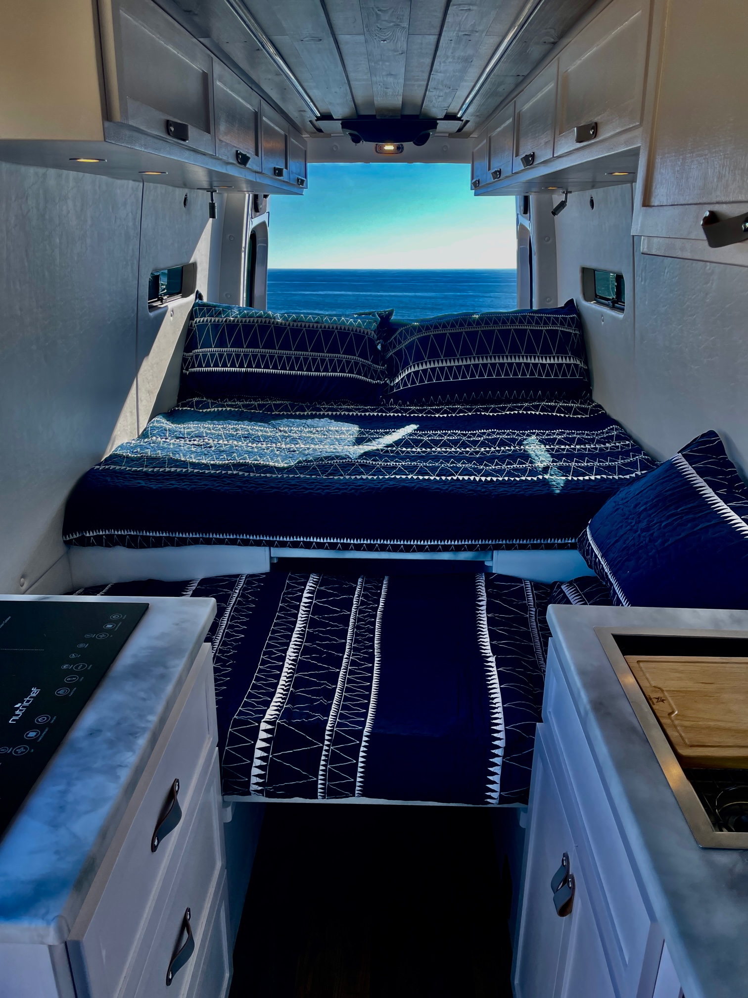 Step inside this RV rental! A comfy bed with blue bedding faces stunning ocean views, with a compact kitchen in the foreground.. Mercedes-Benz Sprinter 2022