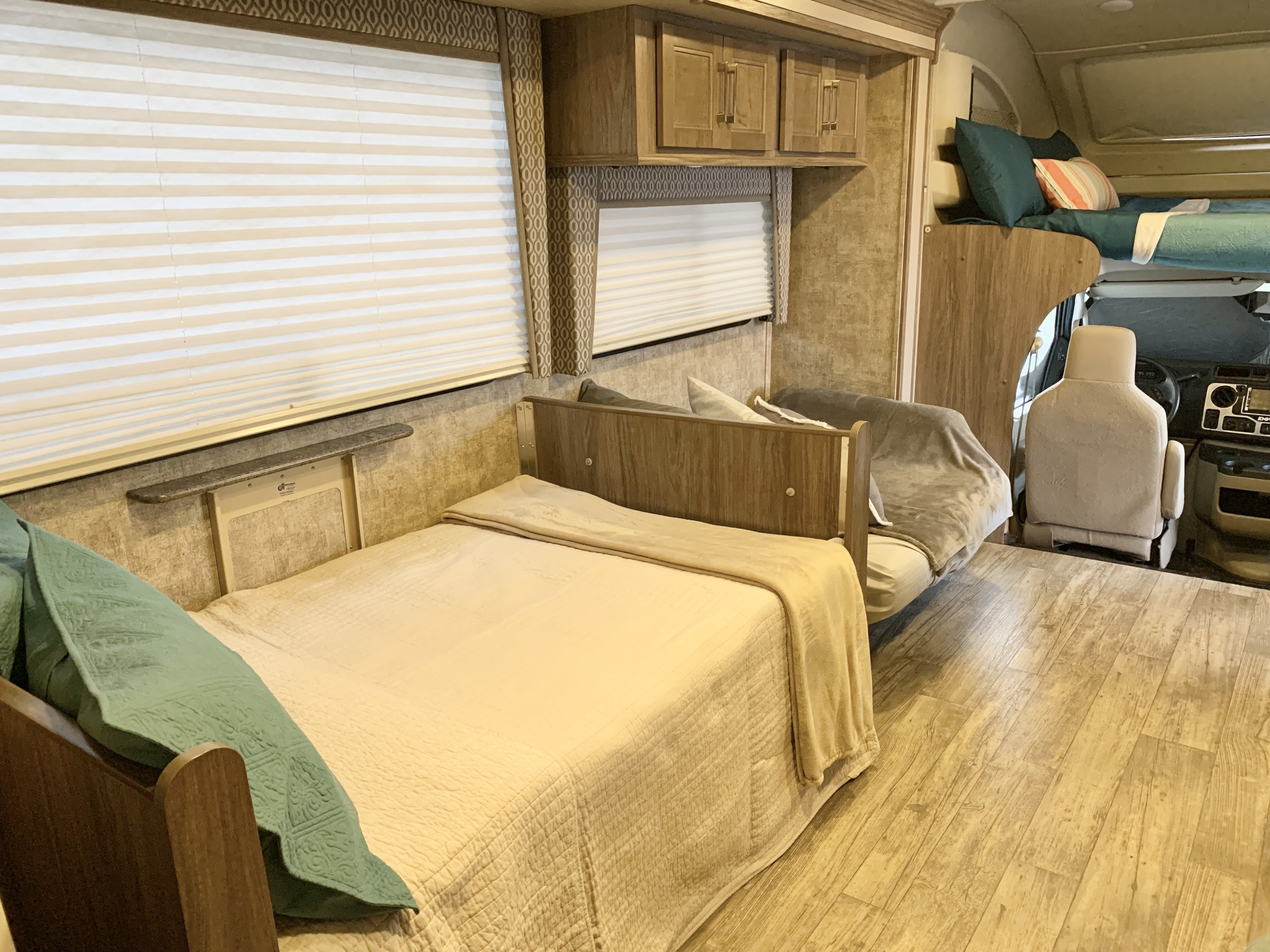 Inside an RV rental, showing the main bed with teal accents, a sofa, bunk bed, and light wood flooring for comfortable travel.. Jayco Envoy 100 2018