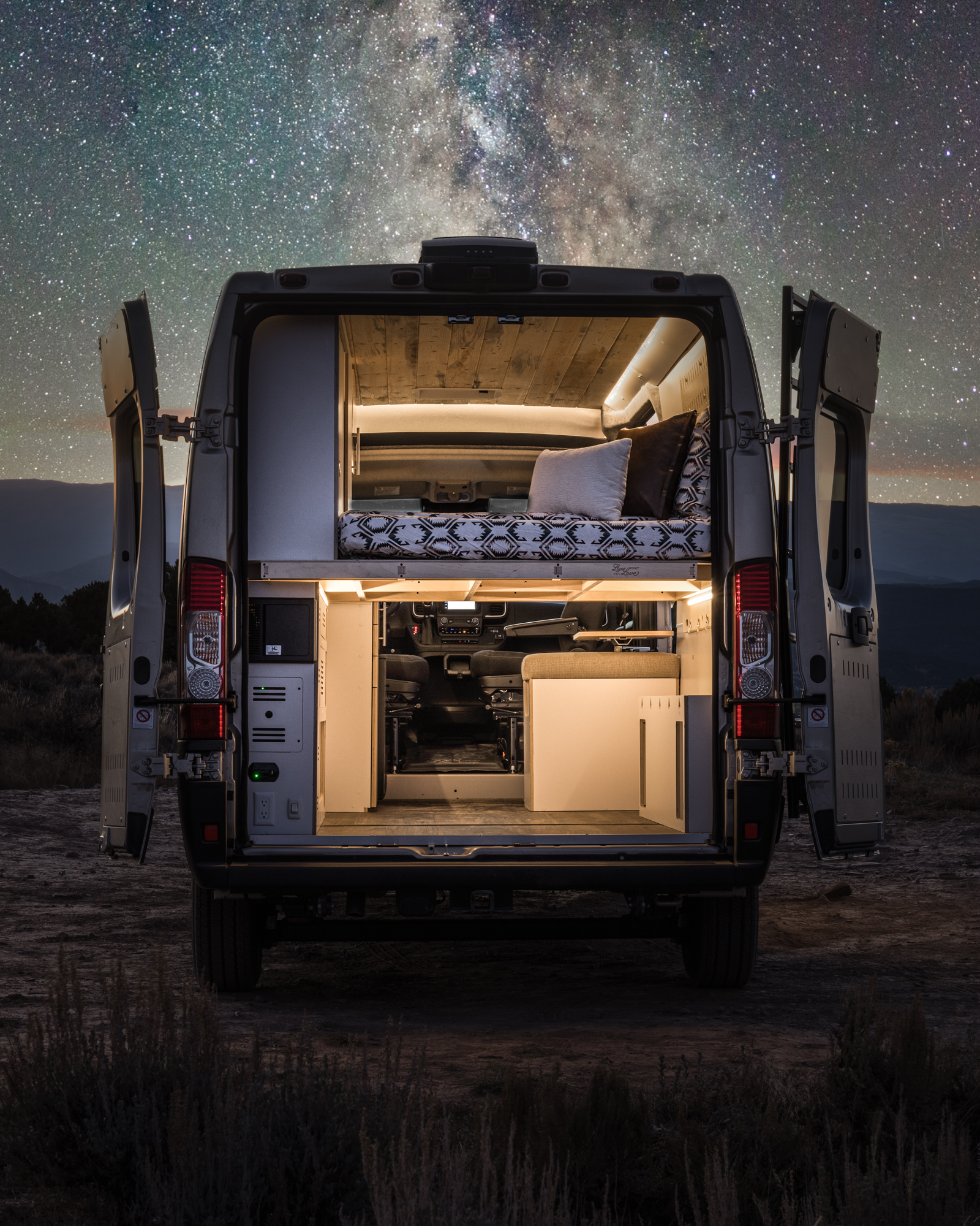 Experience adventure with an RV rental! This camper van, with its lit interior and bed, is parked under a stunning starry night sky.. DM Vans LV7 2022