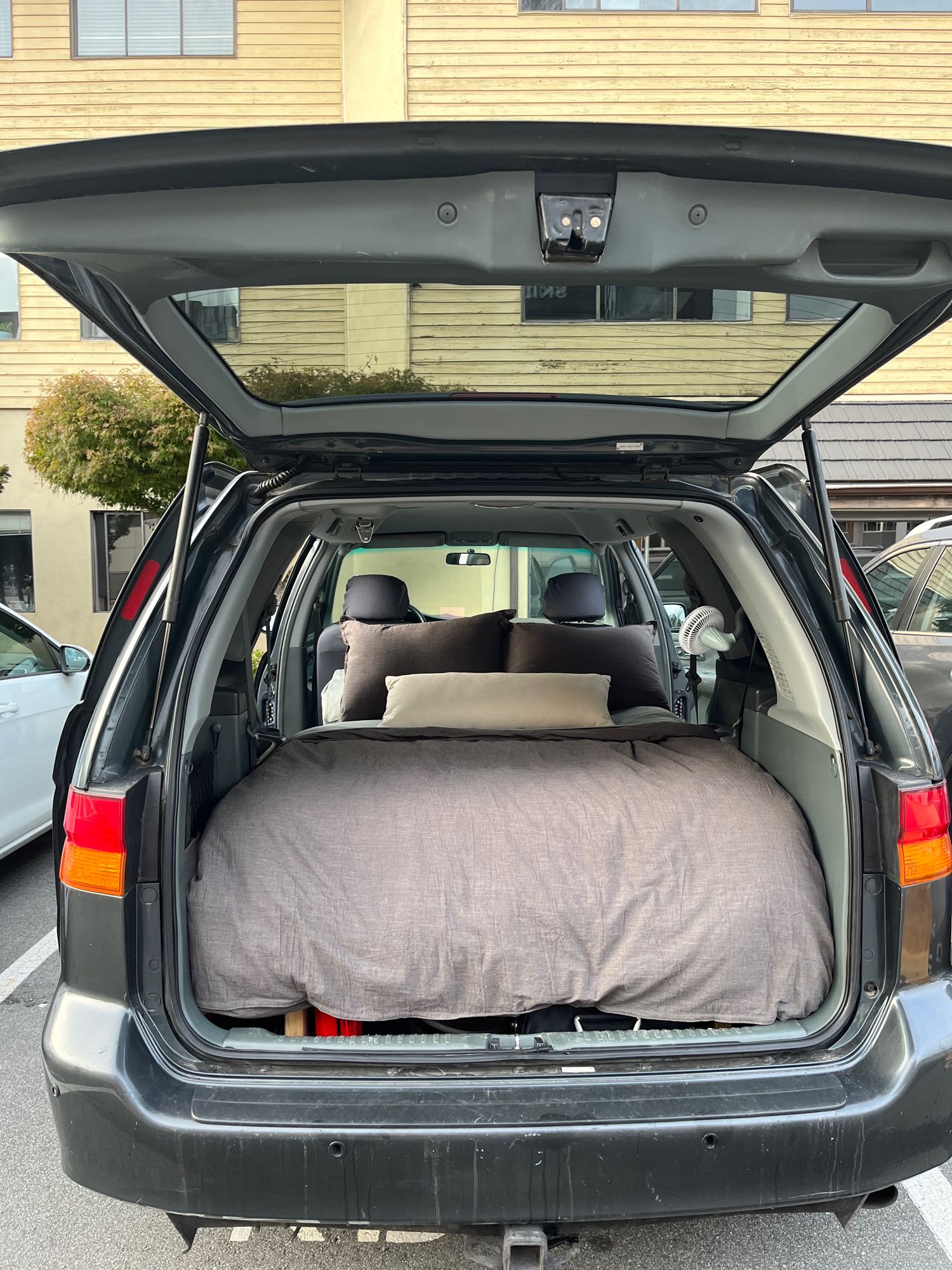 Open SUV trunk shows a bed setup with pillows & fan. A comfy car camping option for road trip travel, akin to an RV rental experience.. Honda Odyssey 2003