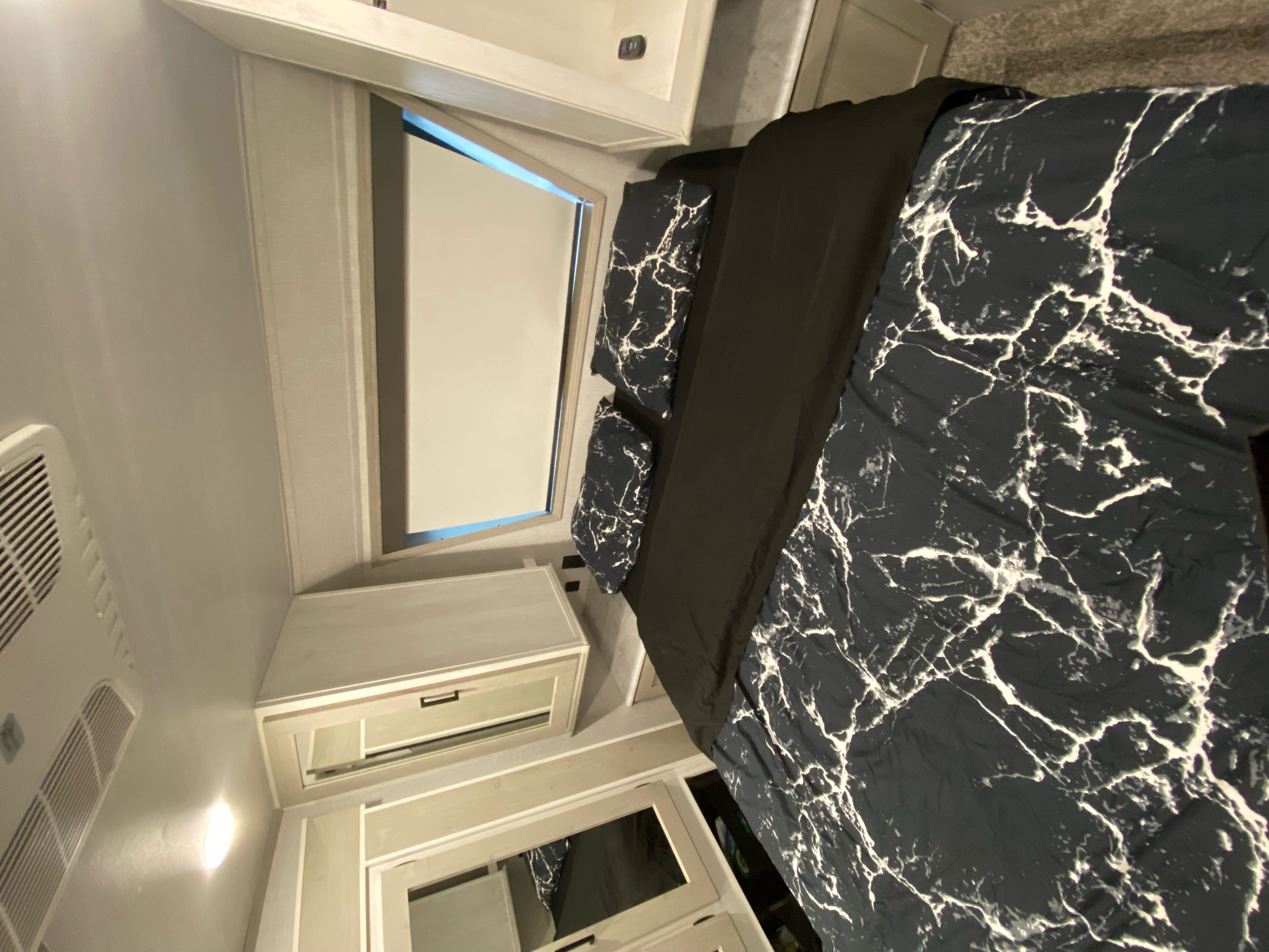 Interior of an RV rental bedroom showcasing a bed with stylish black marble bedding, white storage cabinets, and a pull-down blind on the window.. Forest River Impression 2021