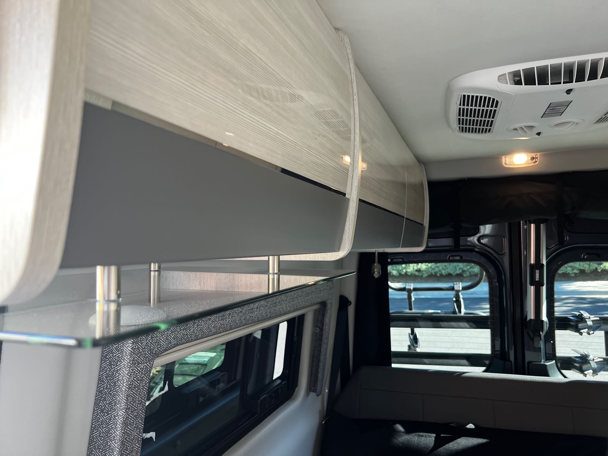 **RV rental** interior: light wood overhead cabinets, ceiling AC, and a clear glass shelf over a window with patterned trim.. Thor Motor Coach Tranquility 19P 2023