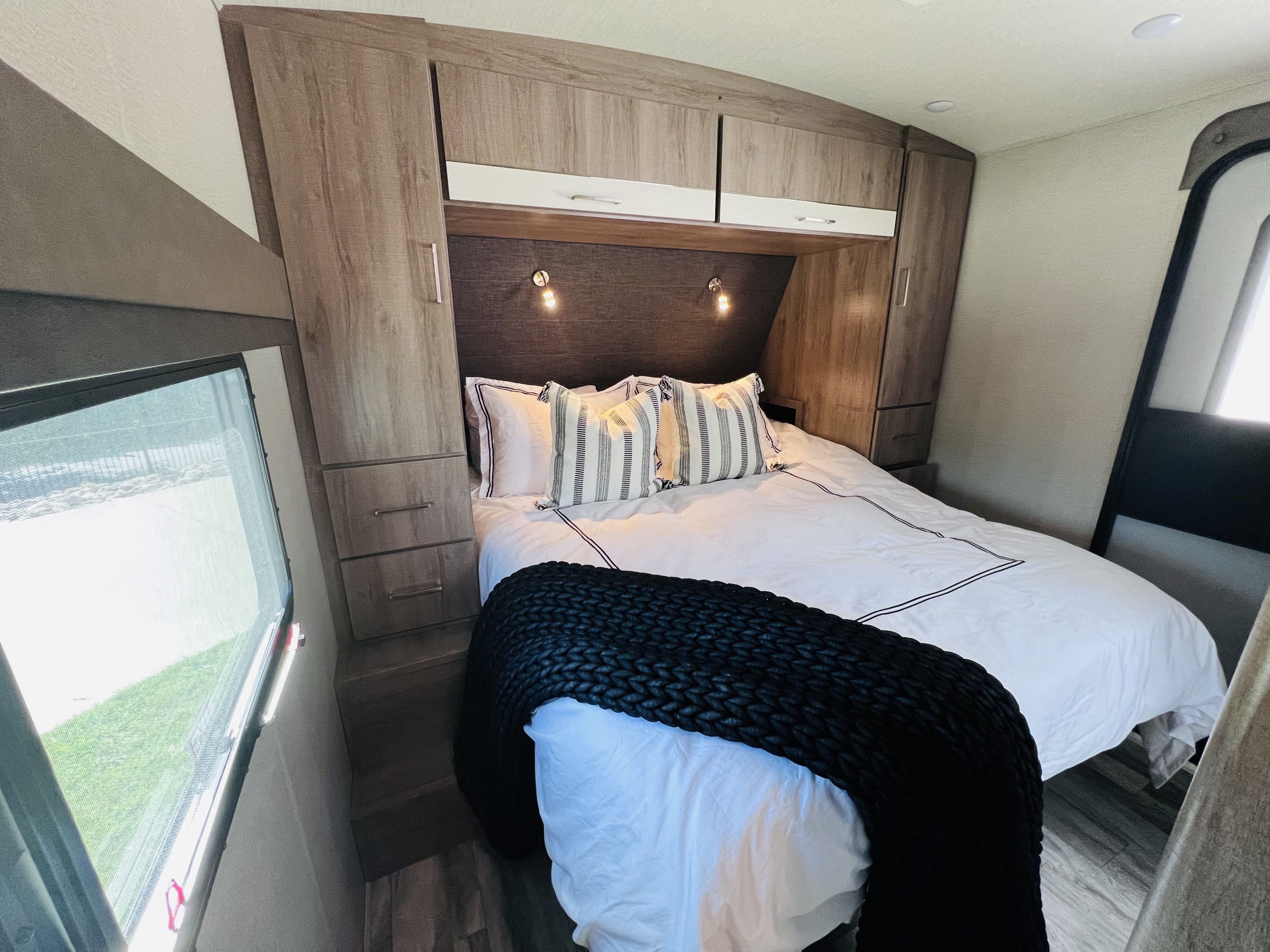 RV rental bedroom: a comfy bed with white linens, sleek overhead cabinets, and a side window, perfect for your next trip.. Grand Design Imagine 2022
