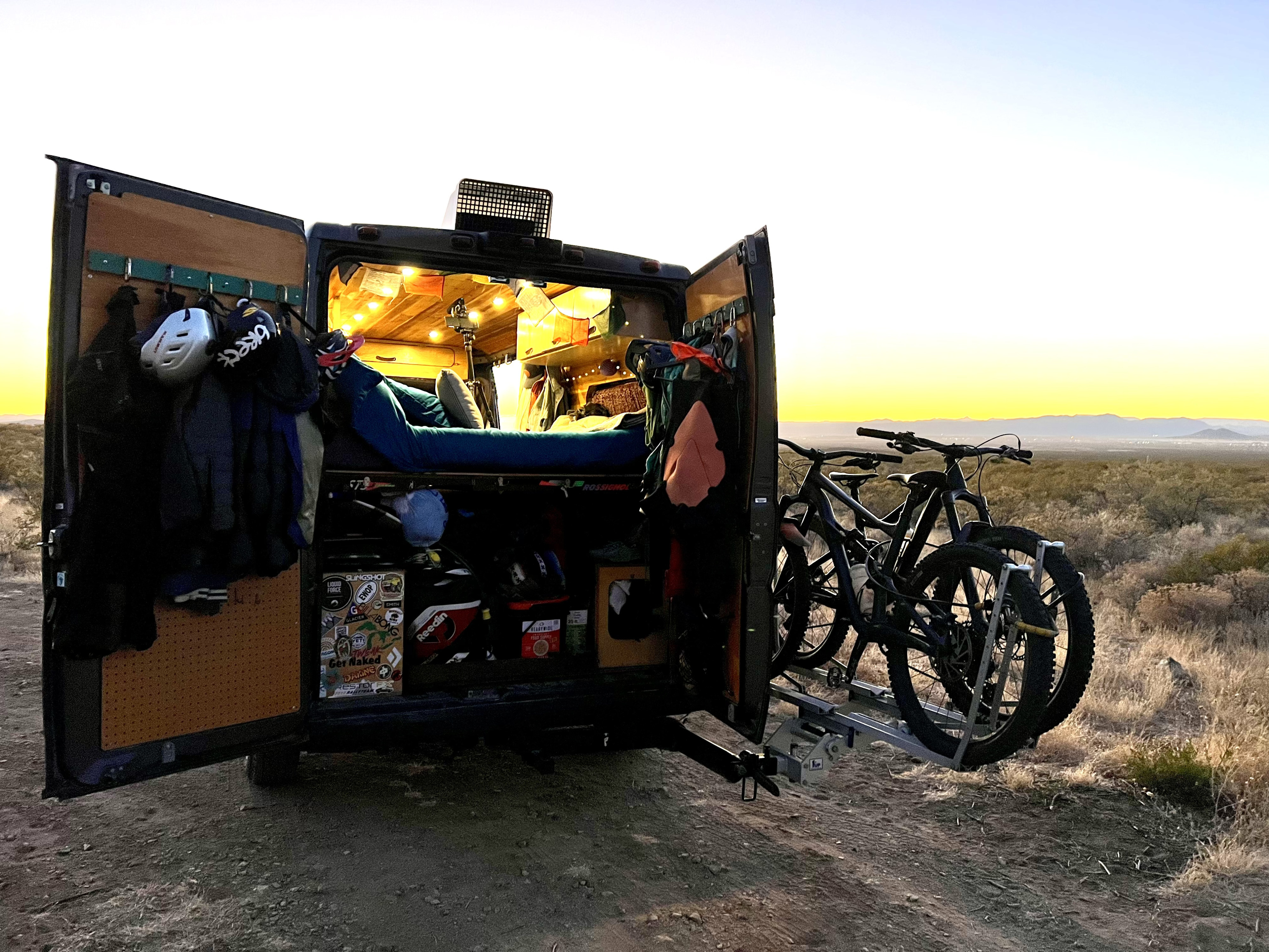 This RV rental camper van, with open rear doors, reveals a cozy interior, bed, gear, and mountain bikes on a hitch rack at sunset.. Dodge Sprinter Van 2019