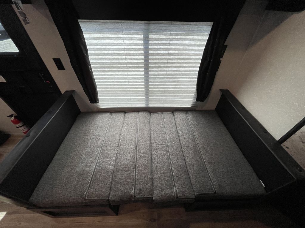 RV rental interior: A gray cushioned bed/dinette area under a window with blinds, flanked by dark curtains. Fire extinguisher on left.. Forest River Cherokee Grey Wolf Special Edition 2022