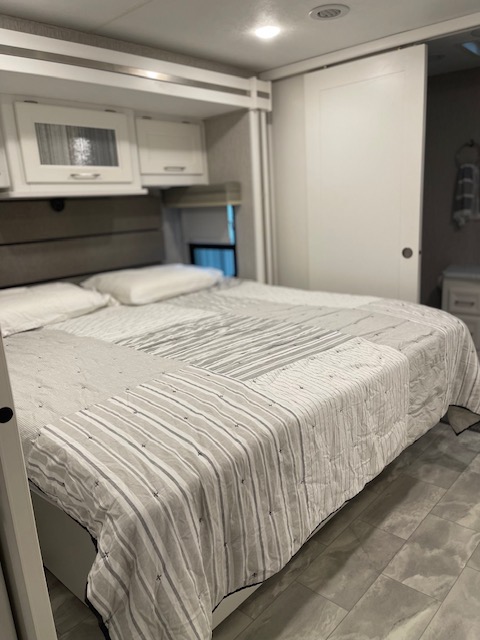 Clean and modern RV rental bedroom featuring a comfy bed with a patterned quilt, overhead cabinets, and a sliding door.. Other Other 2021
