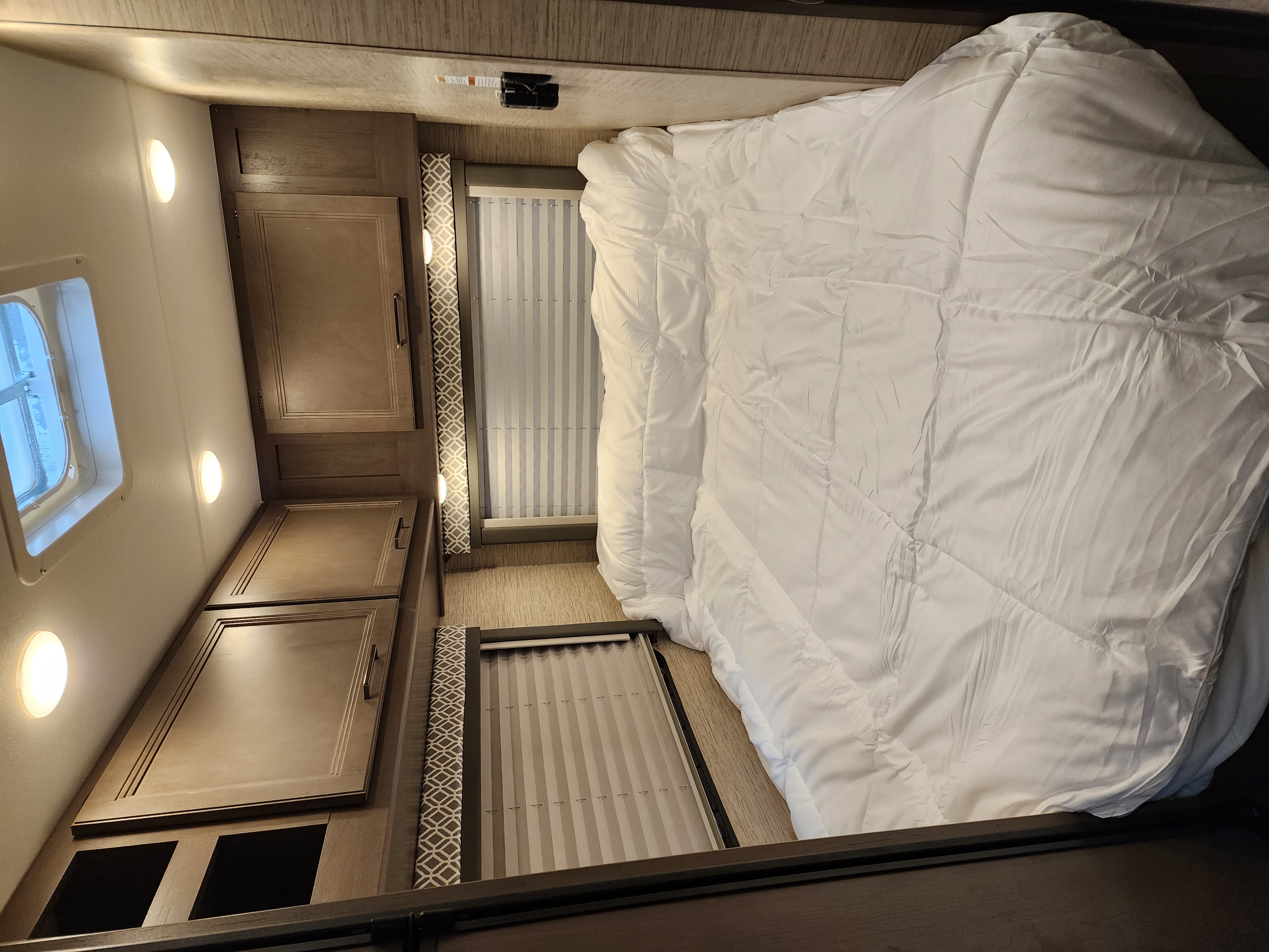 Inside an RV rental: a compact bedroom with a white comforter, overhead wood cabinets, and striped window blinds.. Thor Motor Coach Four Winds 2023