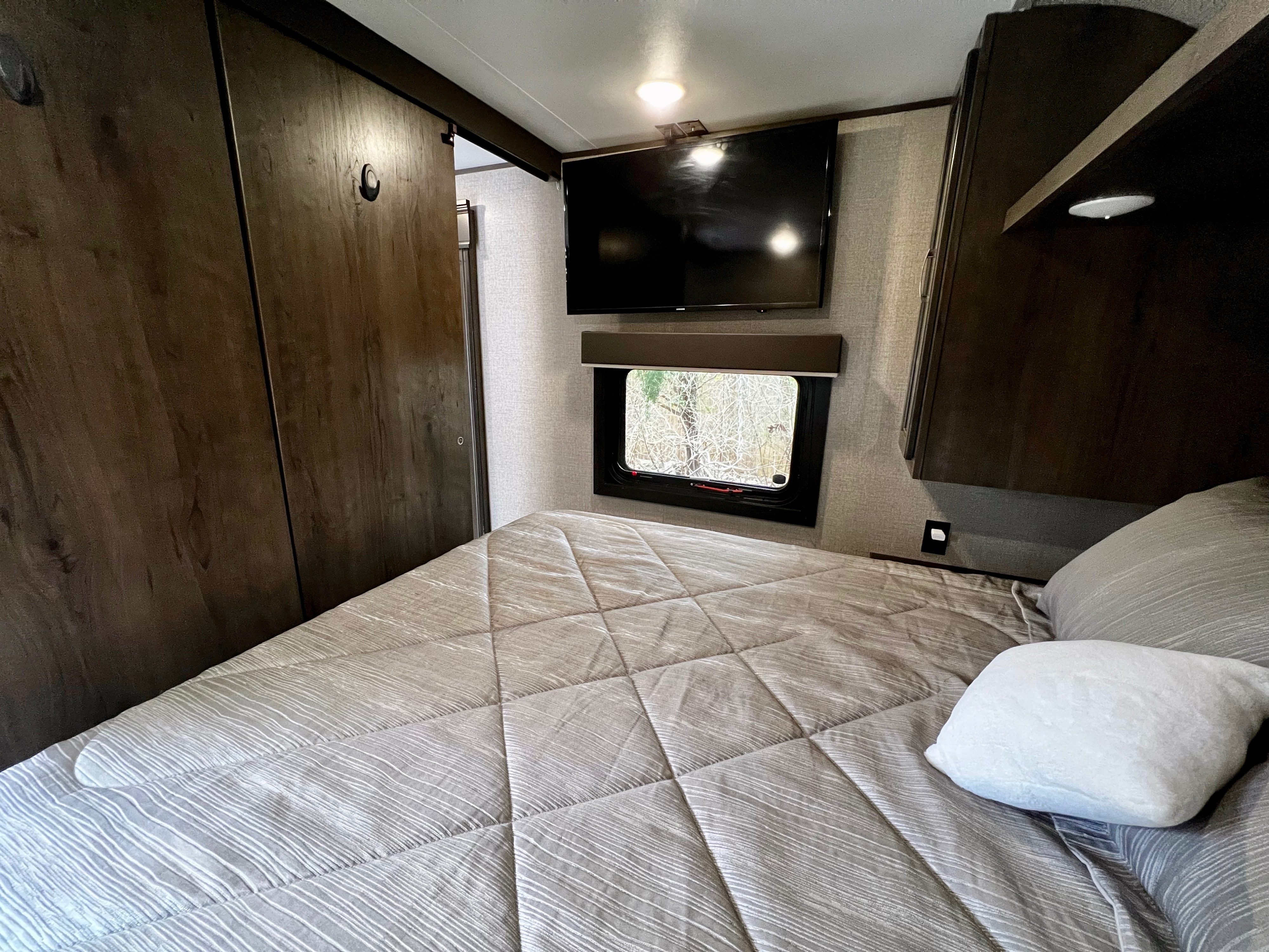 View inside an RV rental bedroom, showing a bed with patterned comforter, wall-mounted TV, and window with outdoor view.. Jayco Jay Flight SLX 2020