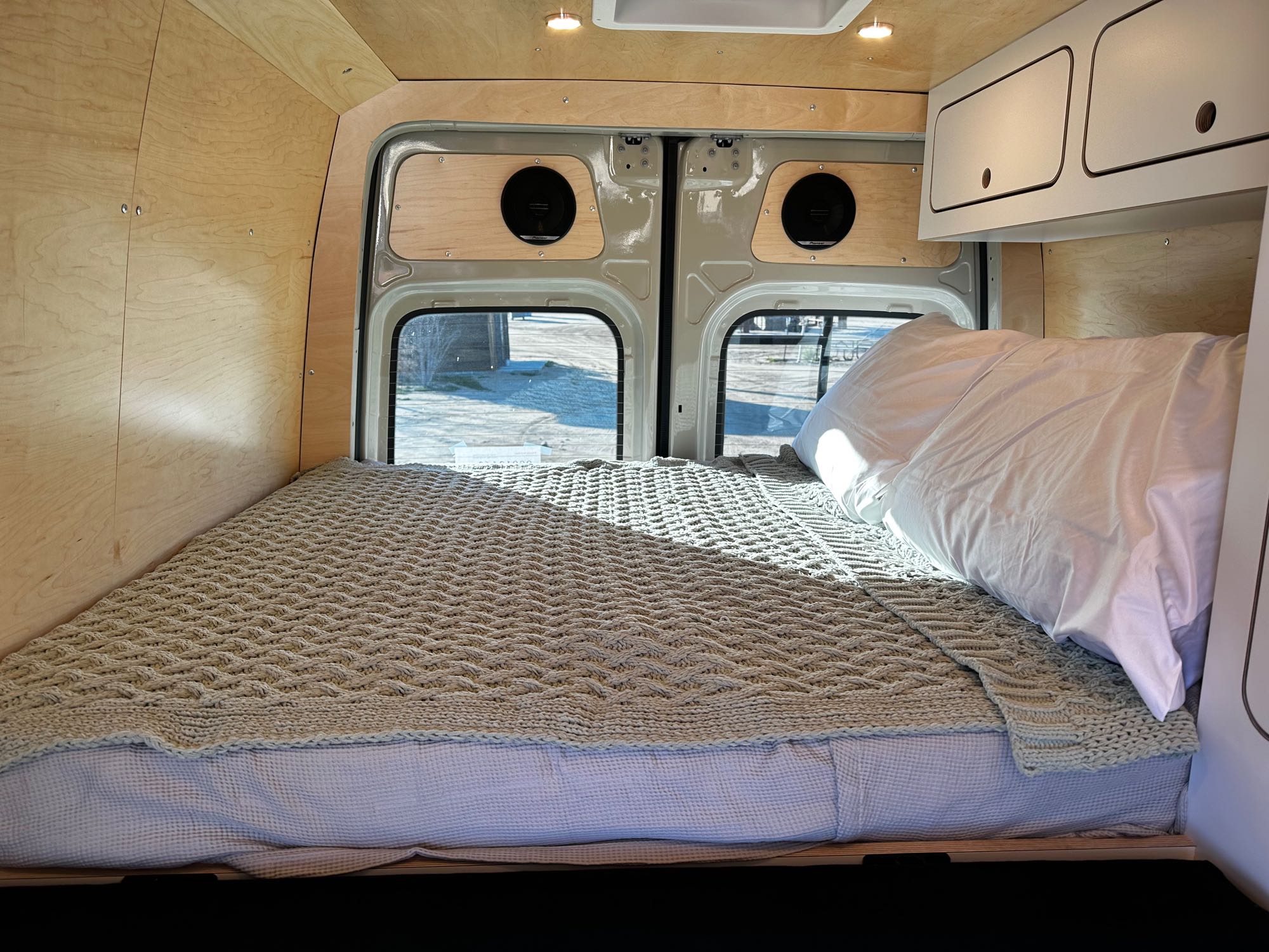 Comfortable RV rental camper van bed with a knitted blanket, wood-paneled interior, and rear windows.. VanCraft VanCraft Class B 2022
